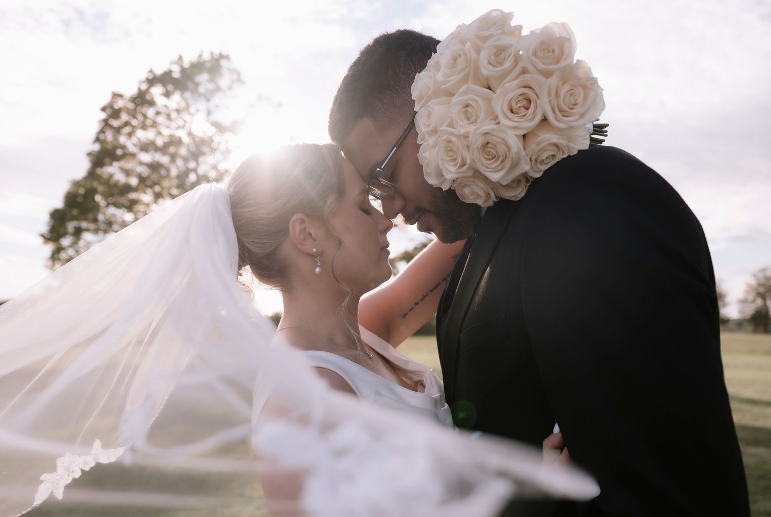 A couple in wedding attire embraces tenderly against a soft white background with pink roses.