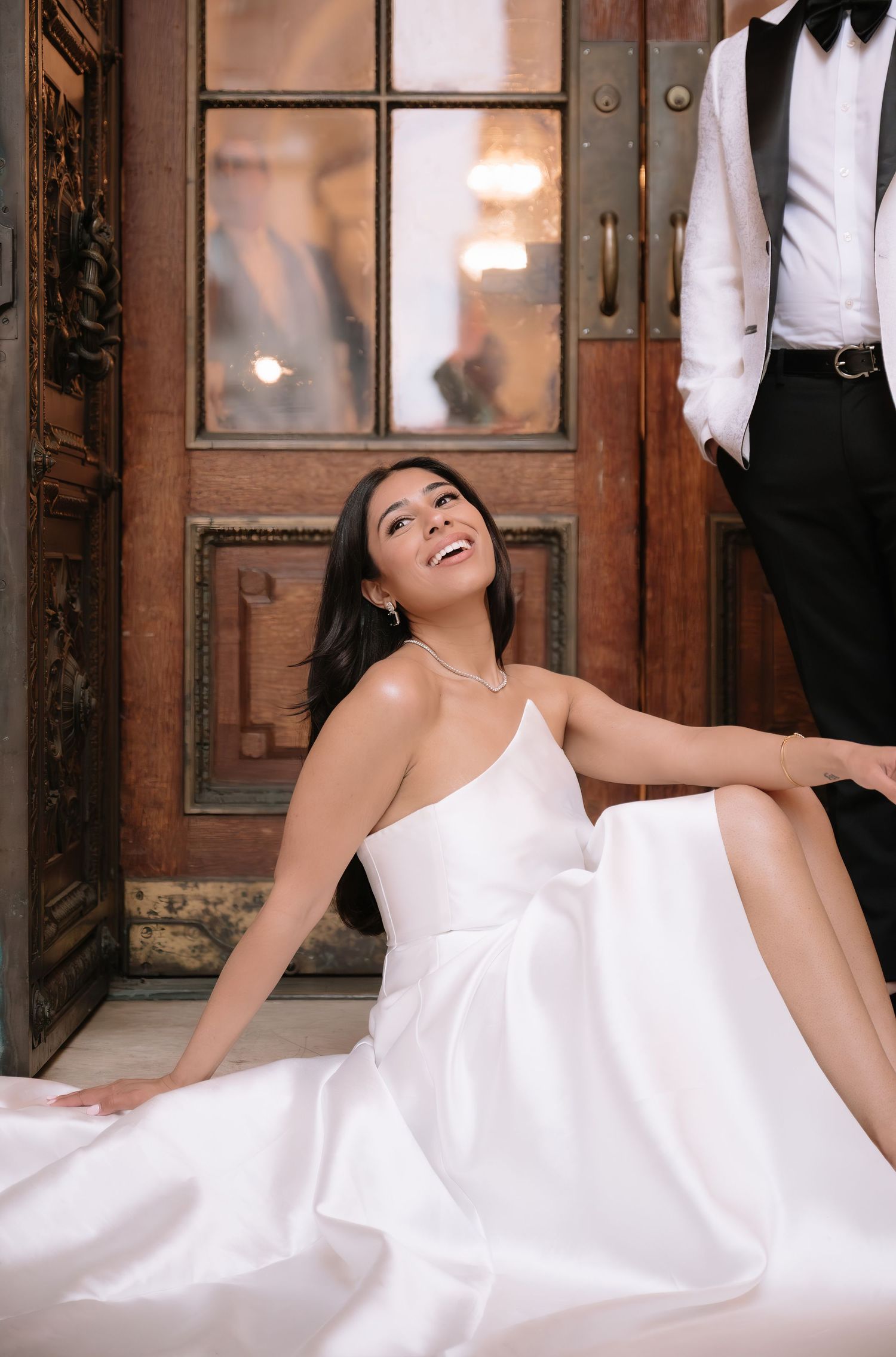 A person in an elegant white gown sits laughing on the floor by a vintage wooden door with an ornate glass panel.