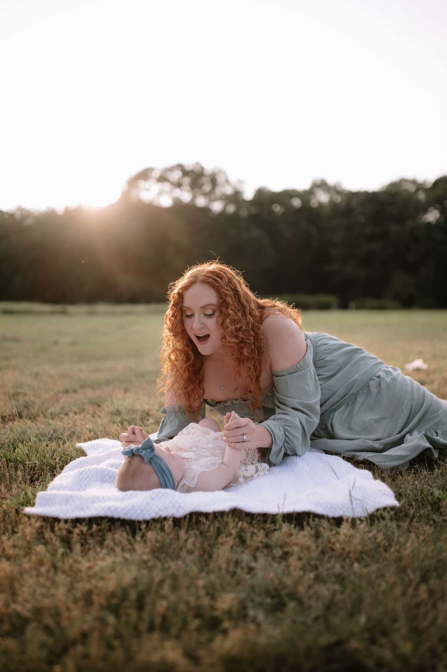 Woman in blue dress lies on blanket in grassy field at sunset with curly red hair glowing in golden light.