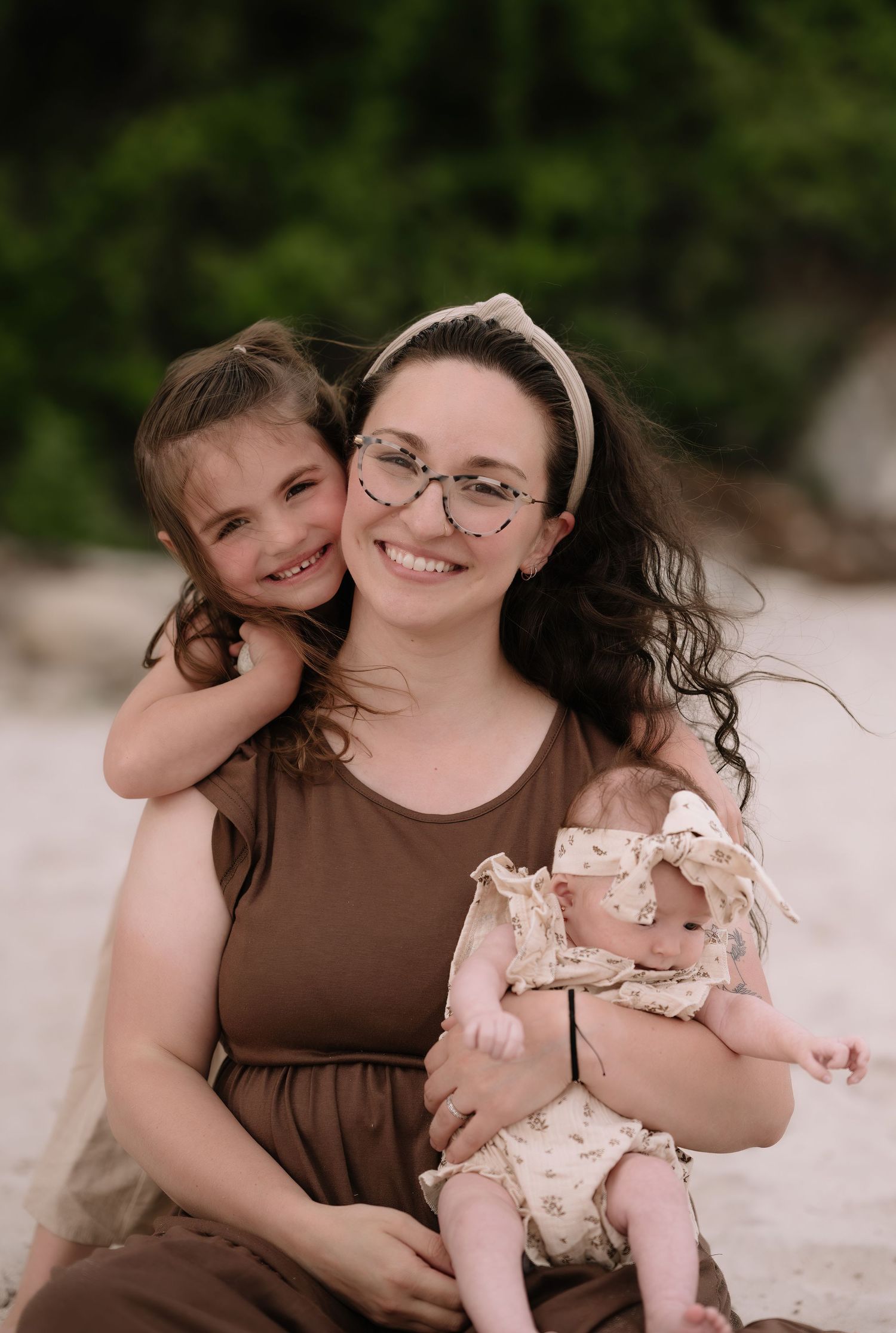 A mother in a brown dress holds her two children while sitting outdoors against a blurred green background.