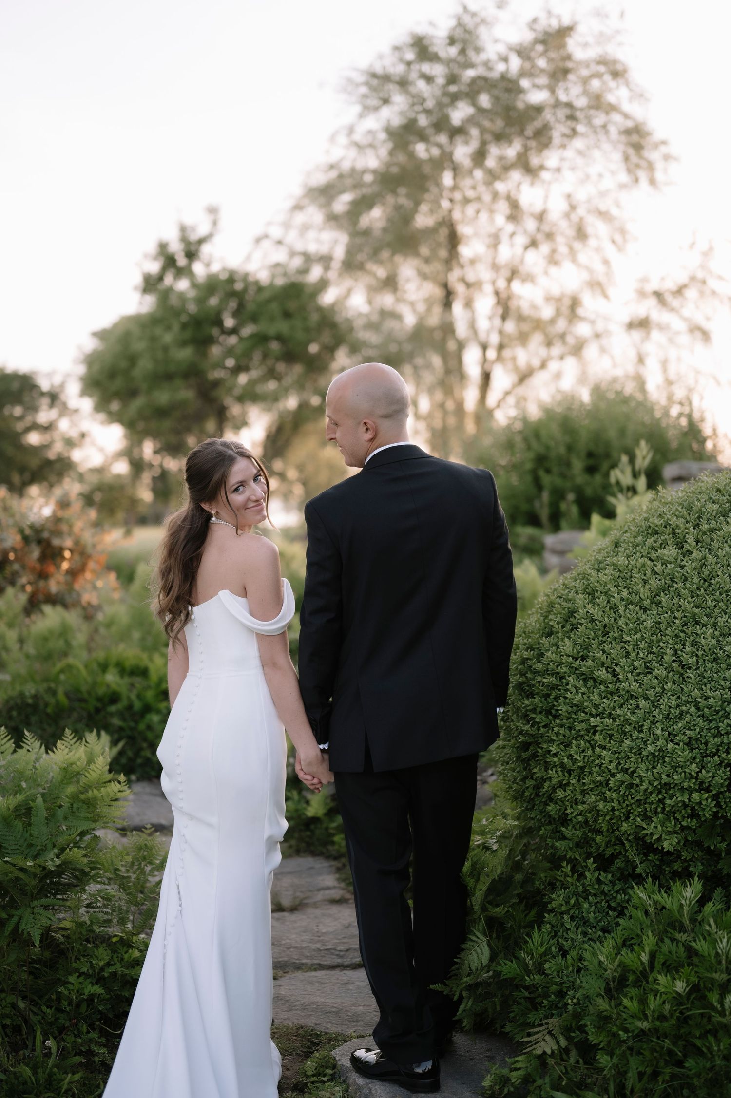 A newlywed couple walks hand in hand through a lush garden pathway during their outdoor wedding.