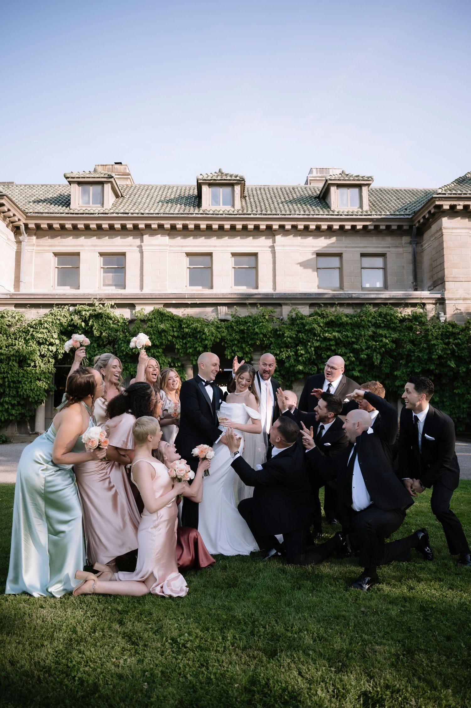 Wedding party gathers together for a fun group photo on the lawn of an elegant historic mansion.