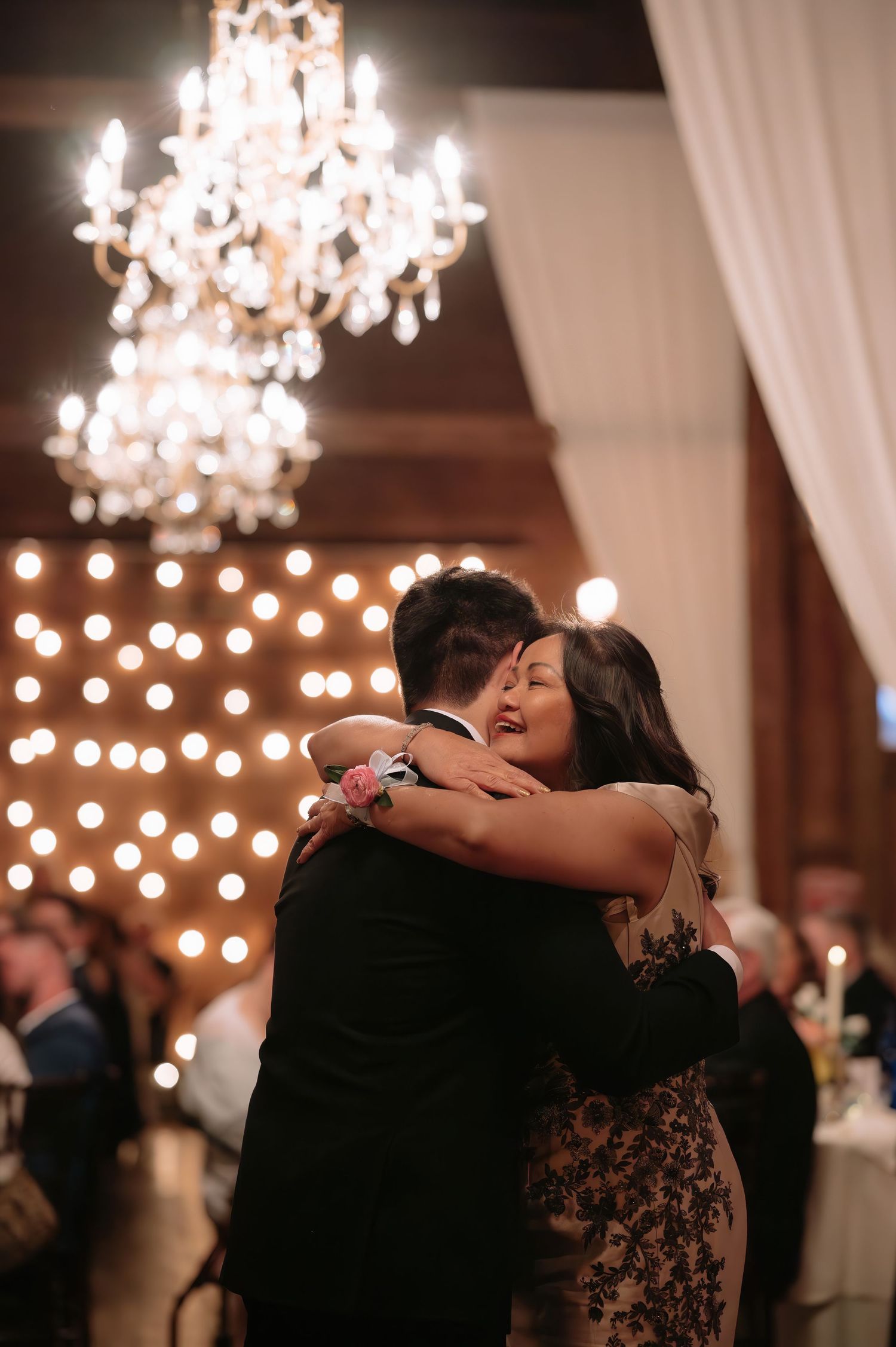 A couple shares an emotional embrace on the dance floor at a wedding reception, with a chandelier and string lights glowing behind them.