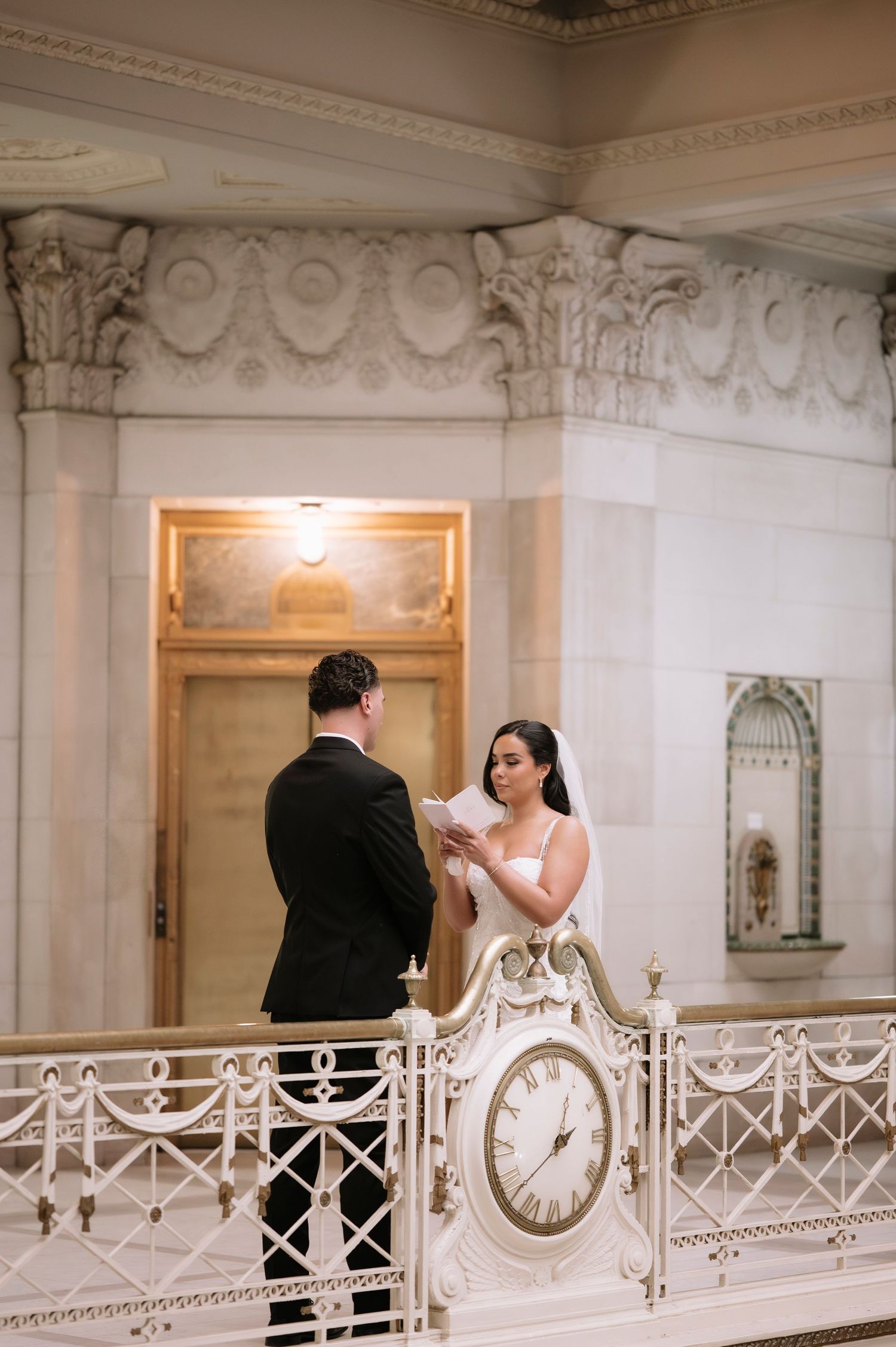 Bride reads vows to groom on elegant balcony with ornate clock at San Francisco City Hall wedding ceremony.
