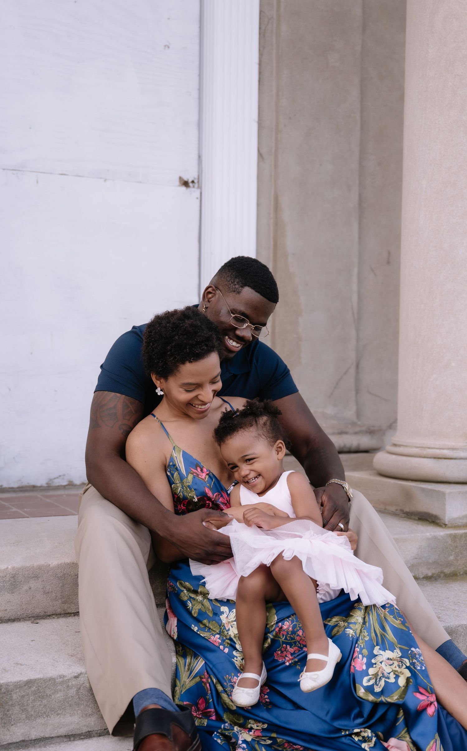 A joyful Black family sits on steps, parents smiling as their toddler daughter laughs in a white dress.