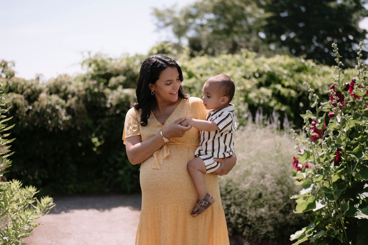 Pregnant woman in yellow dress holds baby in striped outfit in a lush garden path with blooming flowers.