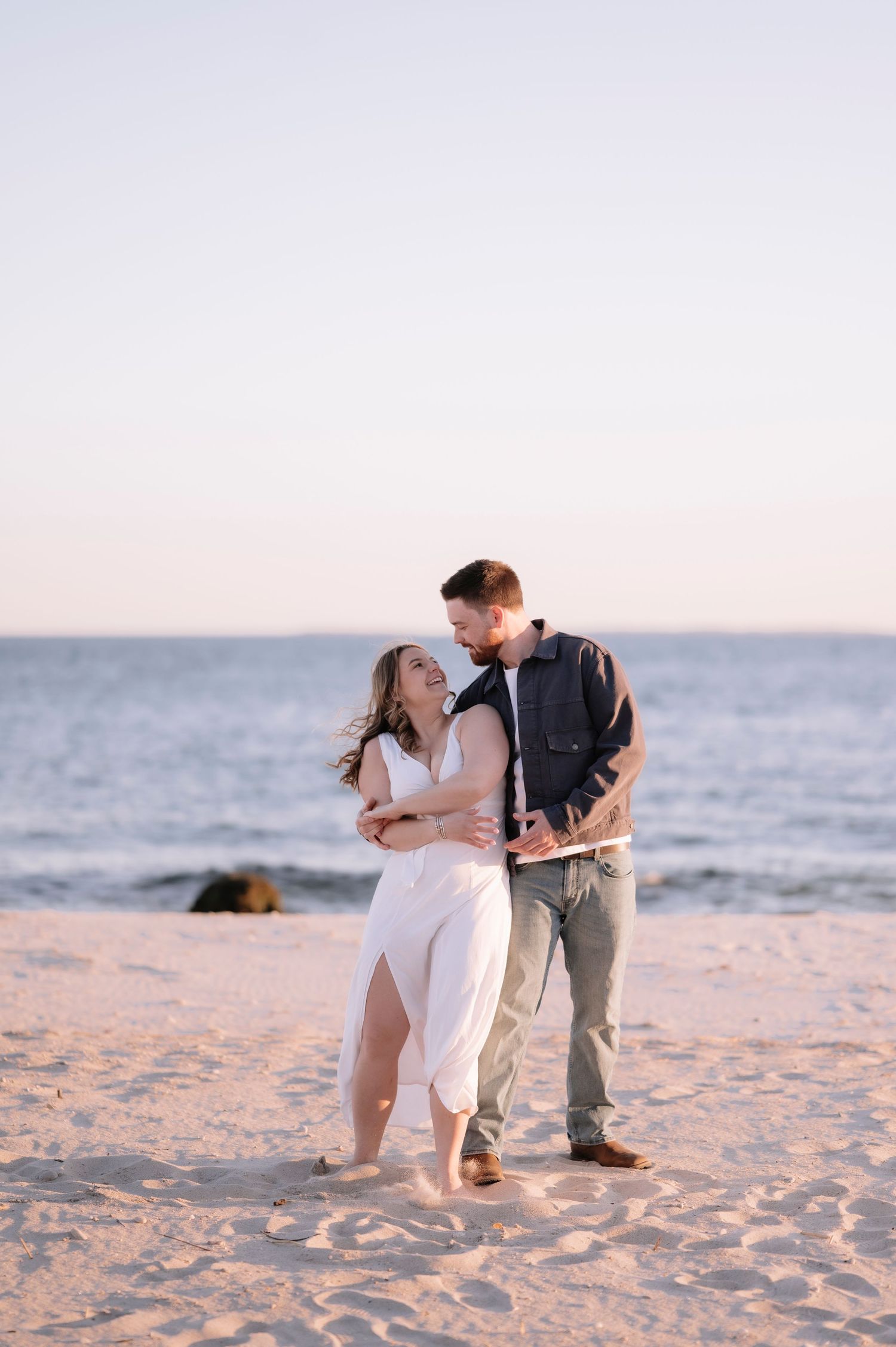 Couple embracing and laughing together on a sandy beach at golden hour with ocean waves in the background.