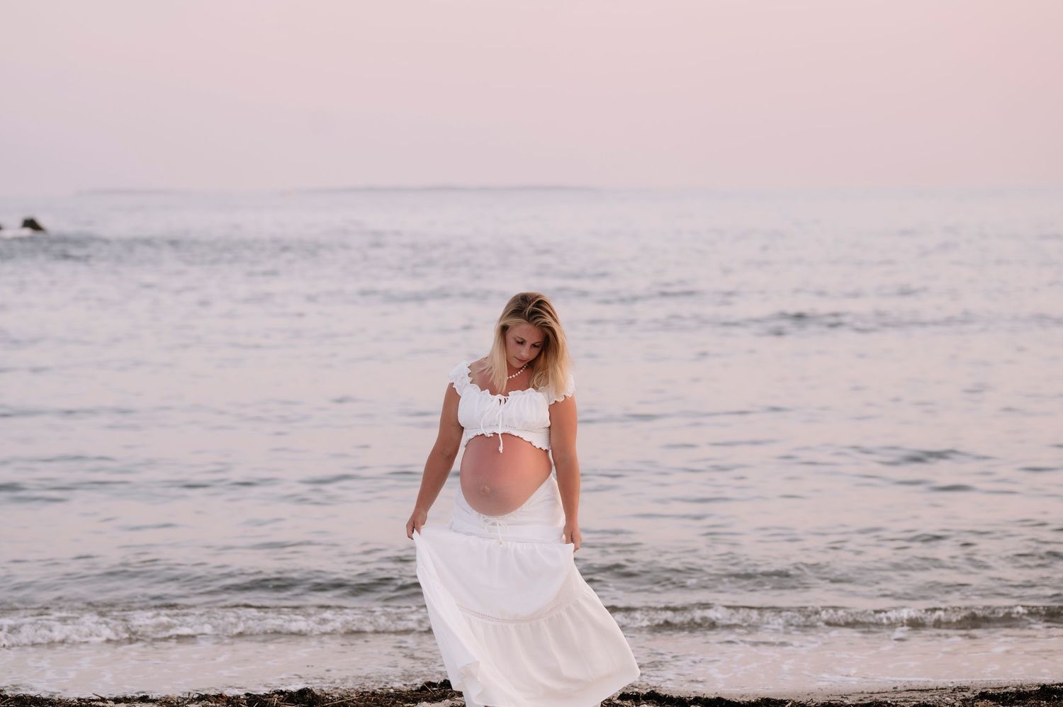 Pregnant woman in white dress standing on beach at sunset, cradling her baby bump by the ocean.