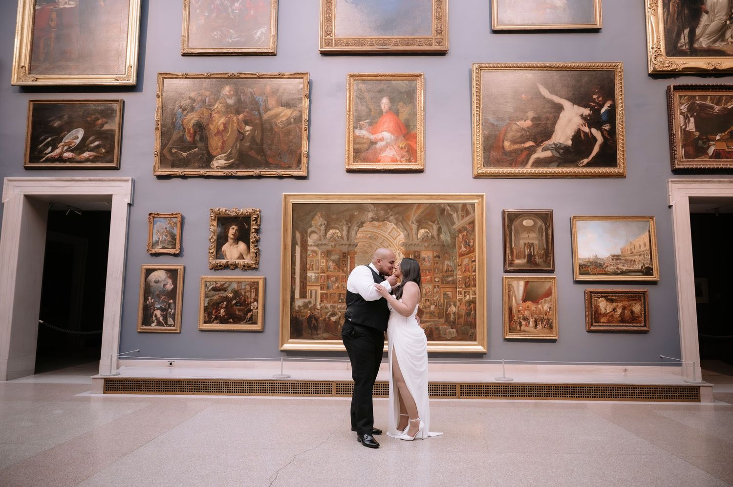 Couple sharing a romantic embrace in an art museum surrounded by large classical paintings on a blue gallery wall.