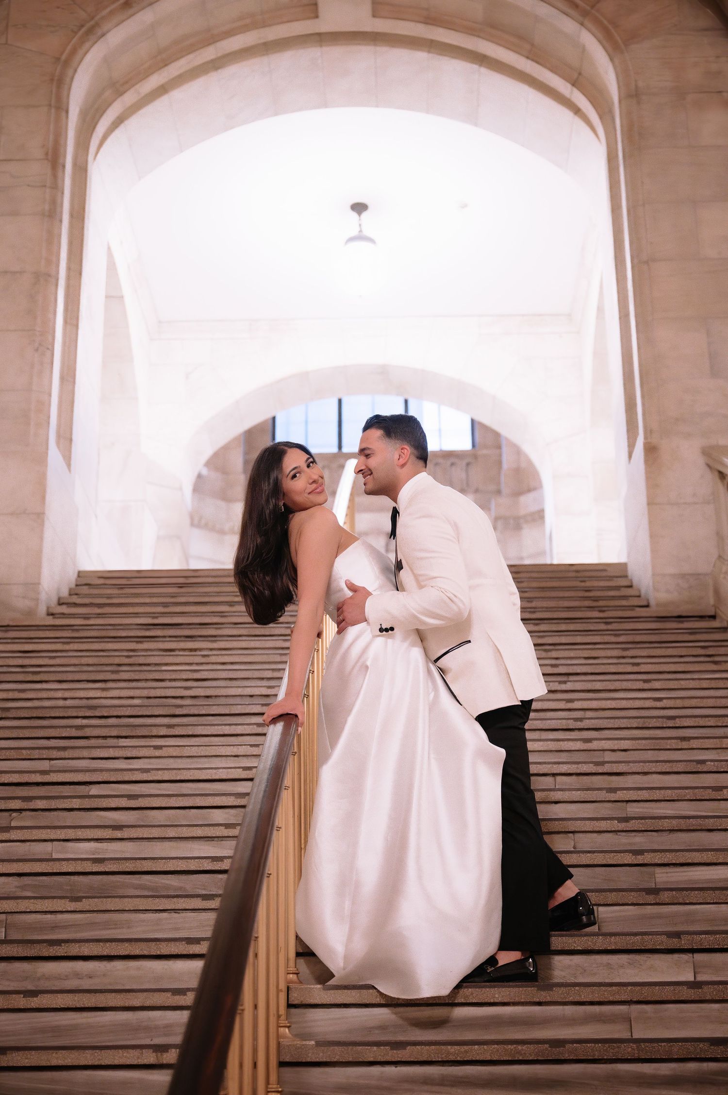 A romantic wedding couple poses on grand stone stairs under an arched ceiling in an elegant architectural setting.