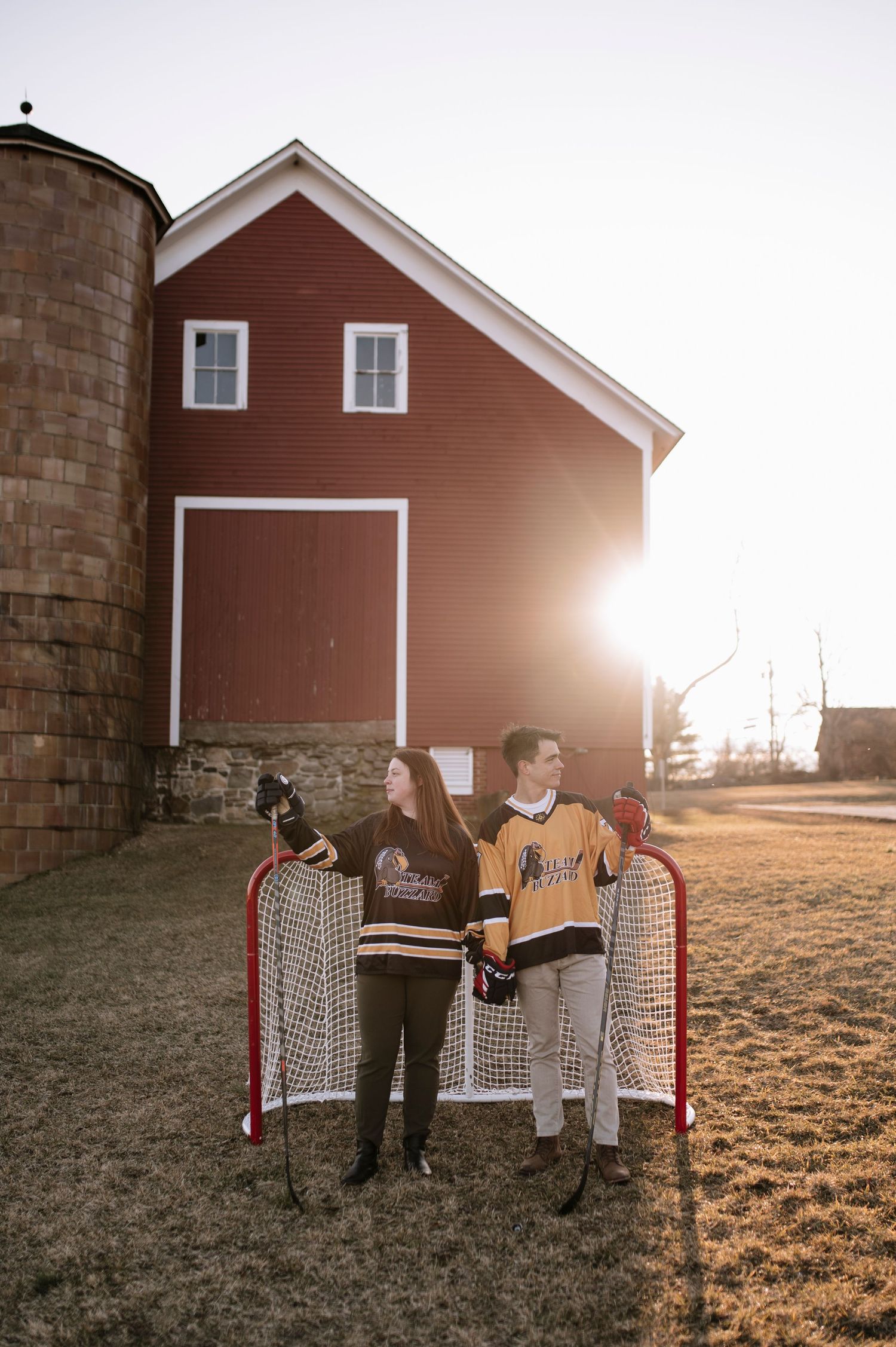 Two hockey fans pose with a goal net outside a red barn at sunset.