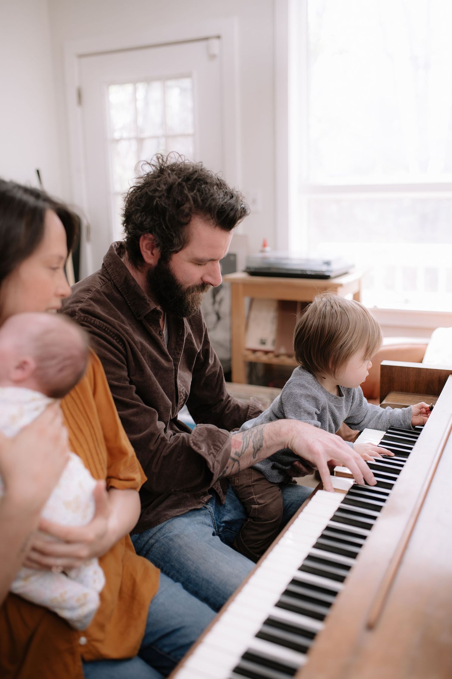 Family gathered around a piano while parent plays, with a baby and toddler joining the musical moment.
