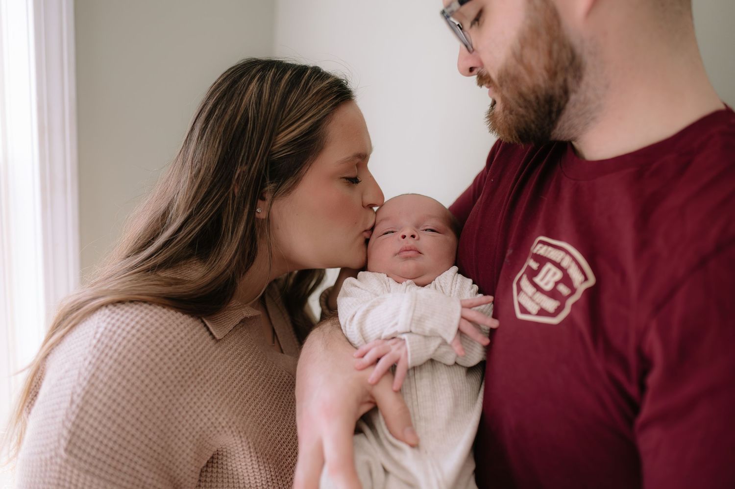 A tender family moment with a newborn baby being held and kissed by parents wearing burgundy and beige.