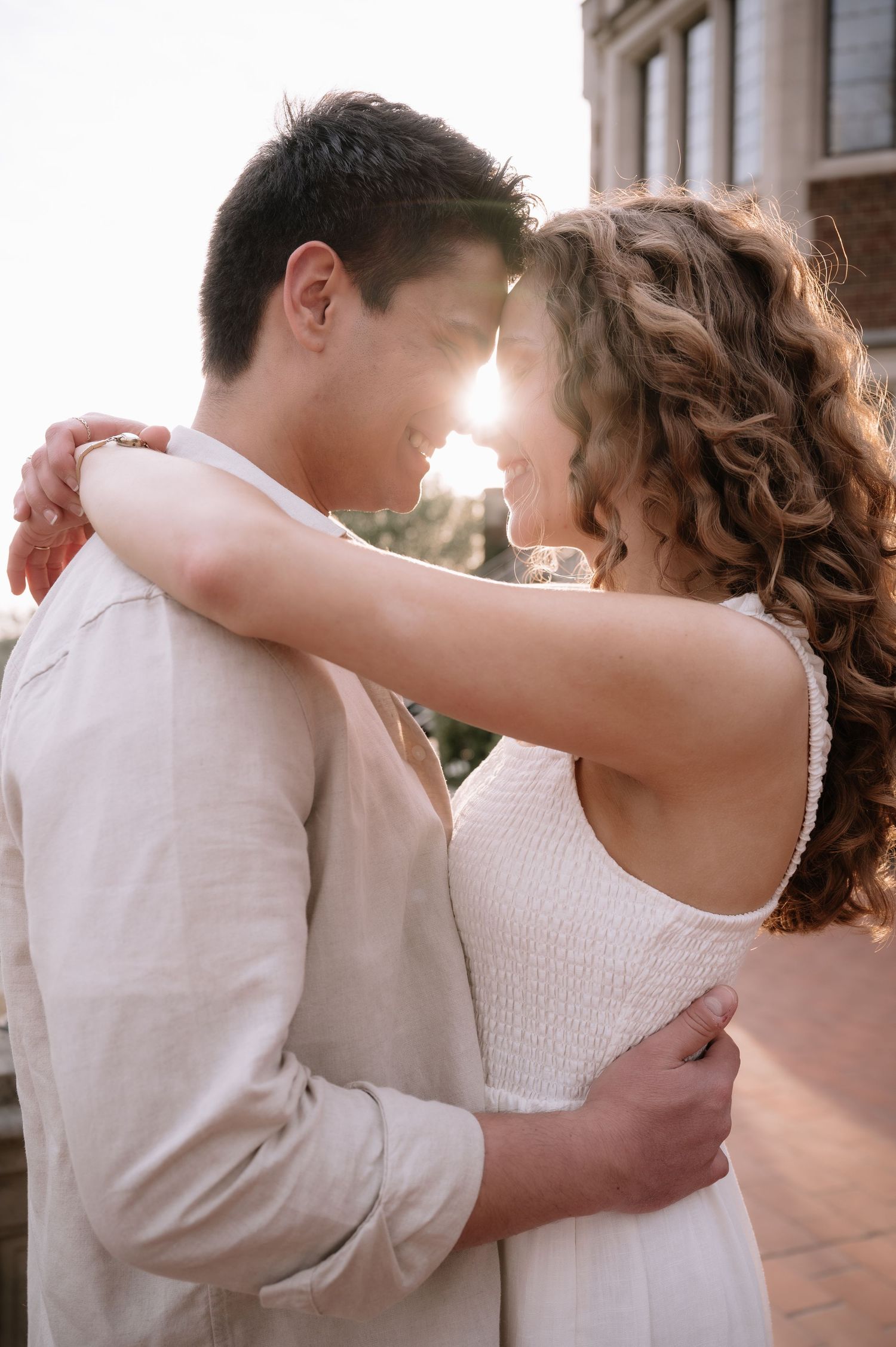 Romantic couple embraces in warm sunlight wearing light colored outfits as sun flares between them during intimate moment.