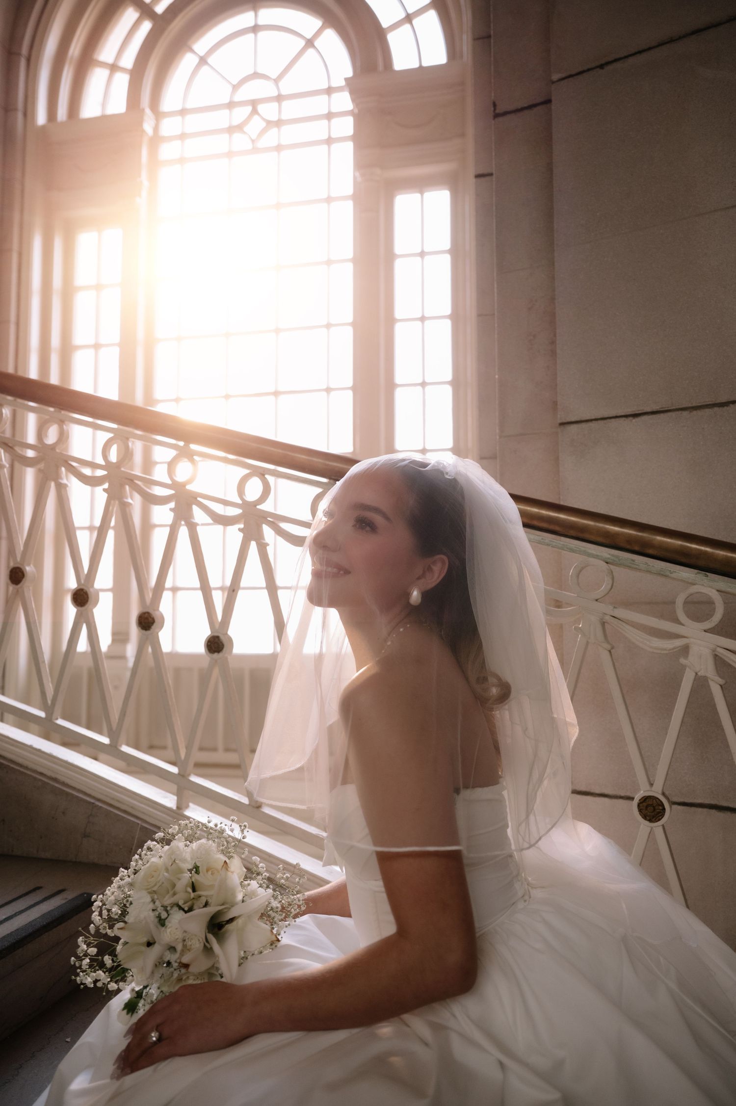 A bride in a white wedding dress and veil sits on elegant stairs near a sunlit arched window holding a white bouquet.