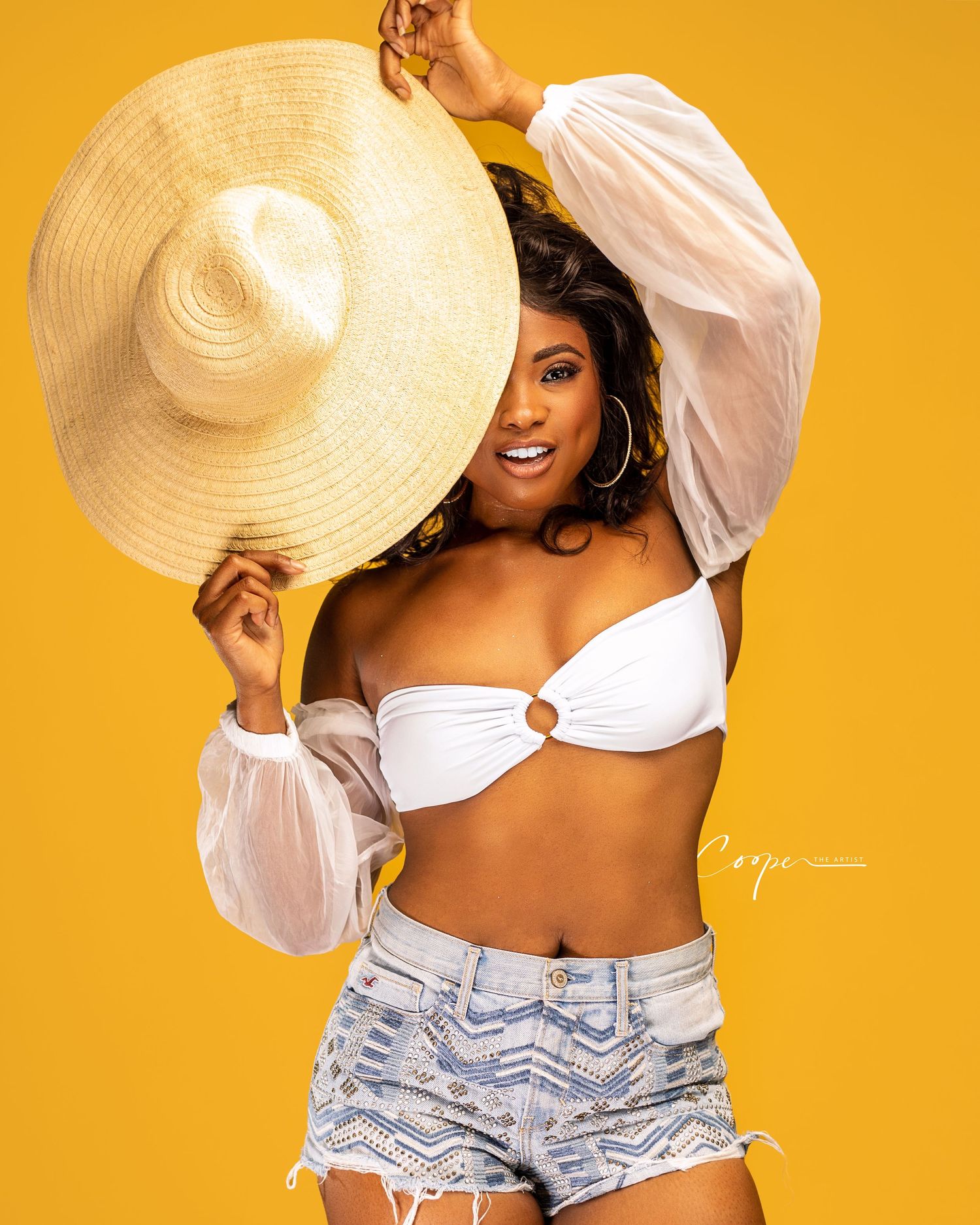 Fashion portrait in white top and denim shorts with large straw hat against yellow backdrop.