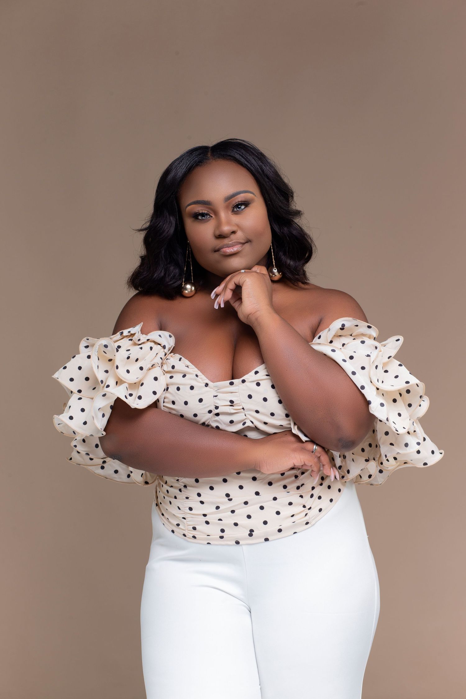 Fashion portrait in polka dot off-shoulder top and white pants against neutral backdrop.