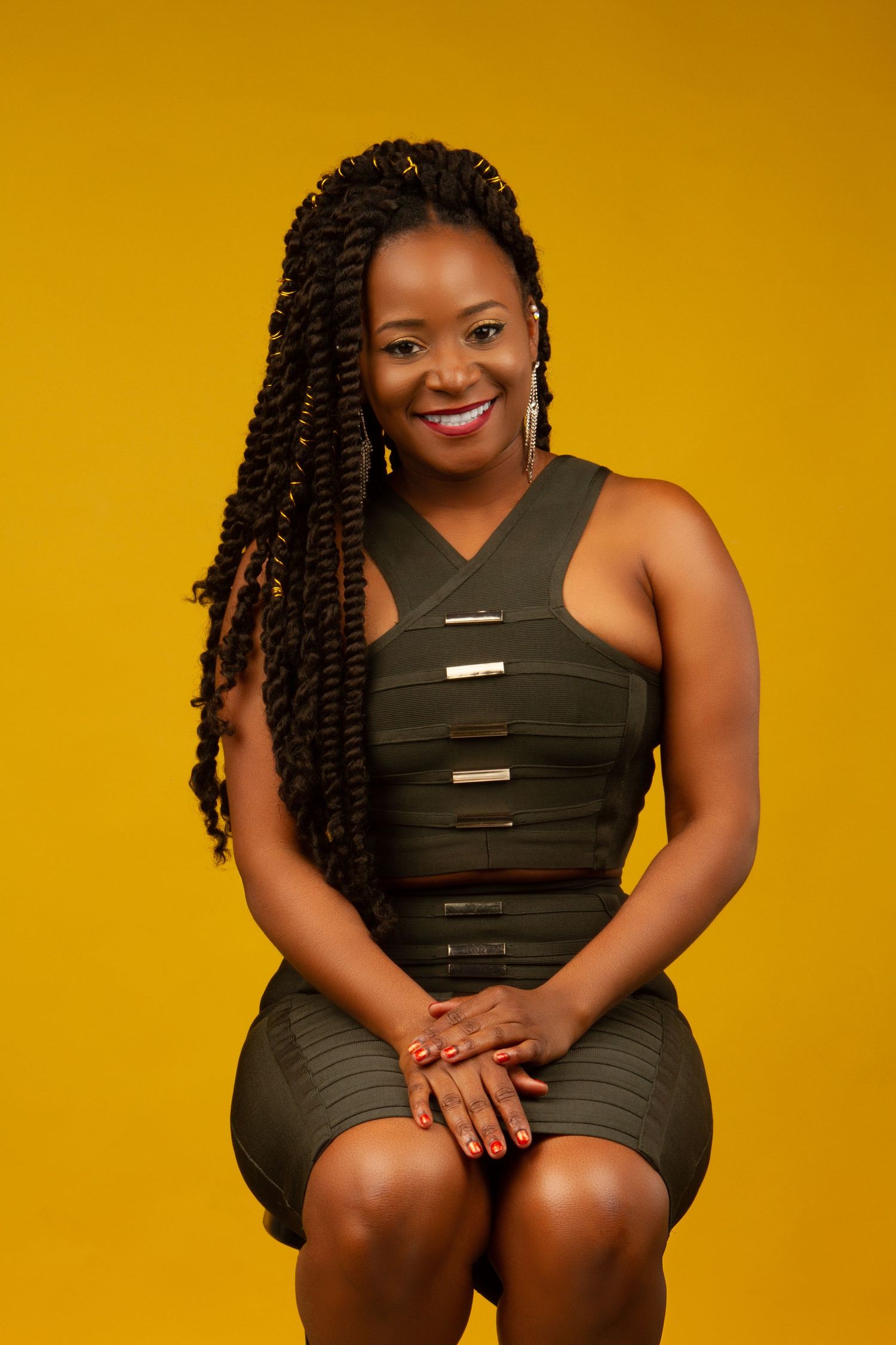 Portrait against yellow background wearing a stylish olive green dress with braided hair and bright smile.