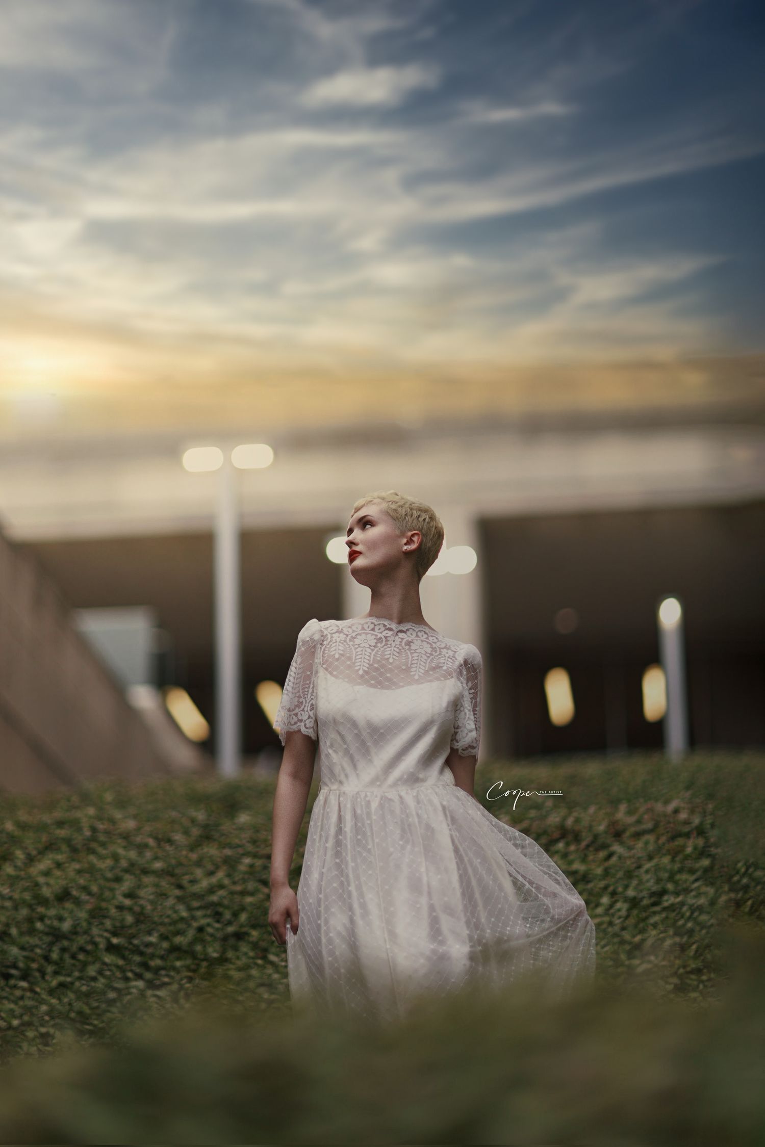 Woman in vintage white dress poses dramatically at sunset in urban setting.