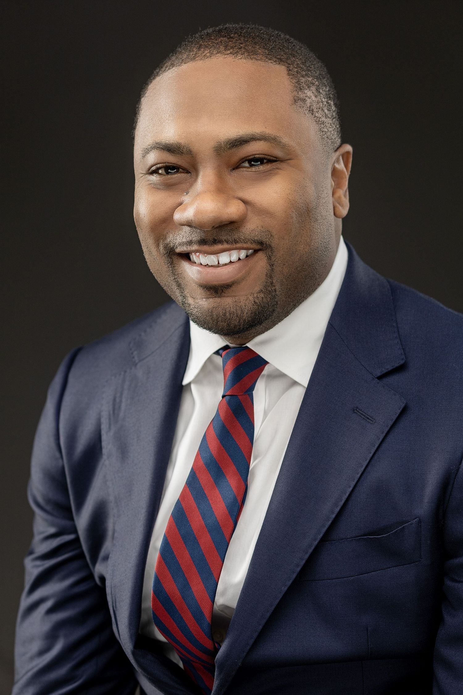 Professional headshot in navy suit with red and blue striped tie.
