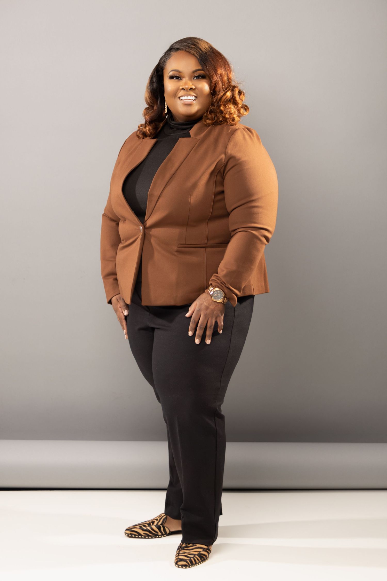 Professional model in brown blazer and black pants strikes a confident pose against gray backdrop.