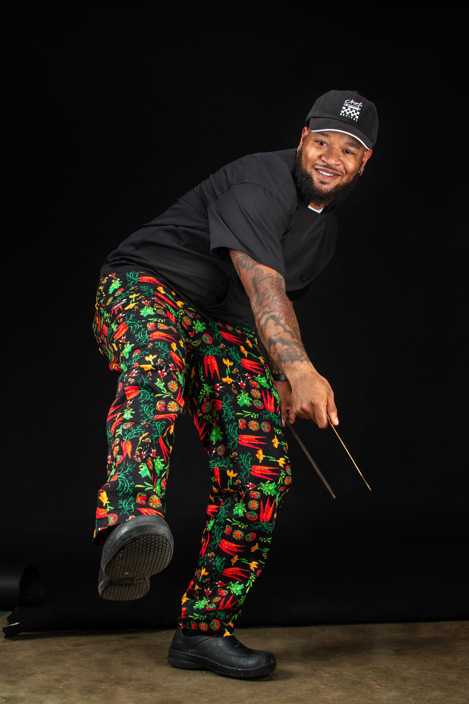 Fun portrait of chef in colorful Christmas-themed pants and black shirt holding drumsticks.