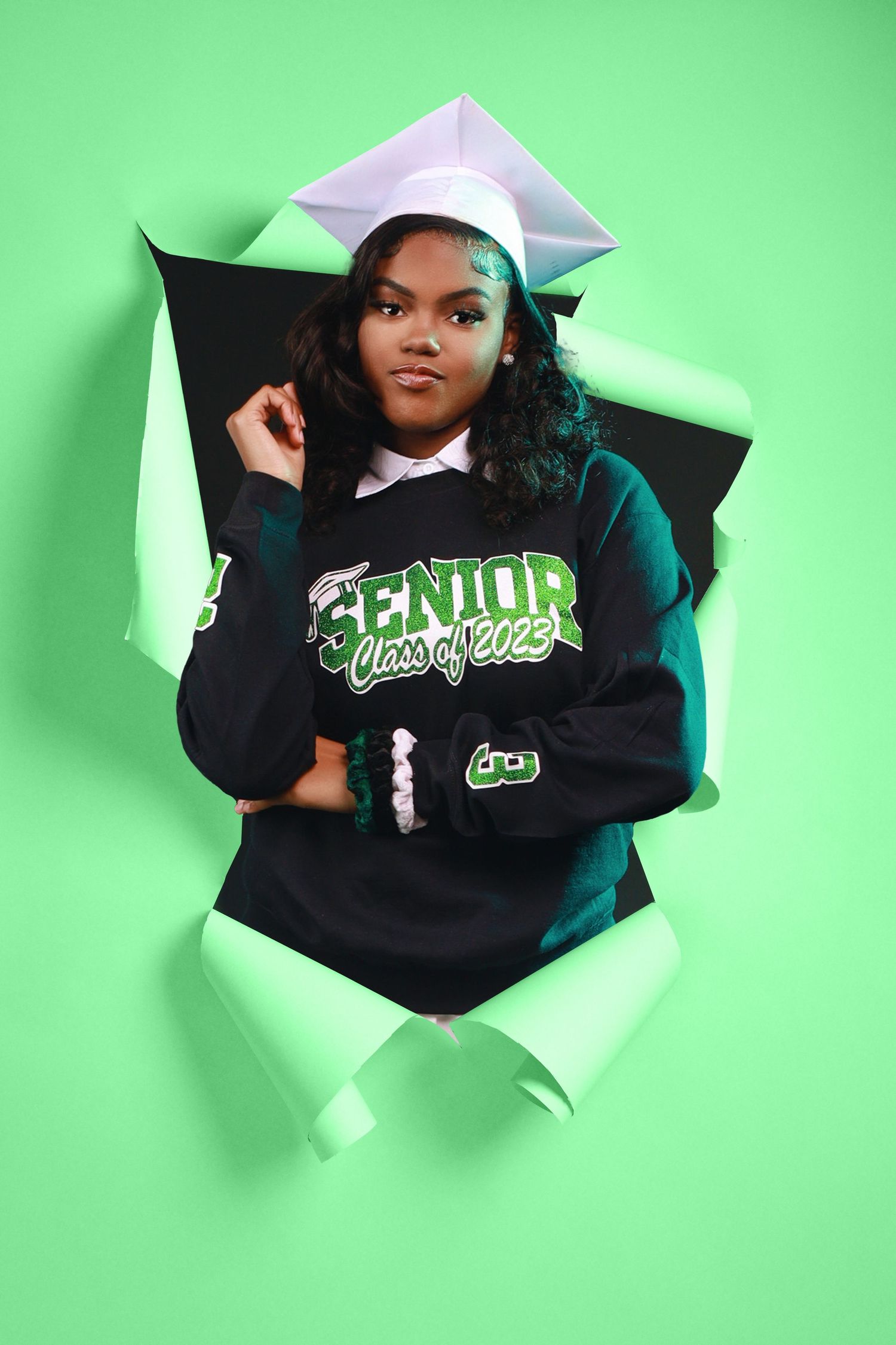 Creative portrait through torn green paper showing graduate in cap and gown wearing 'Senior' varsity sweater.