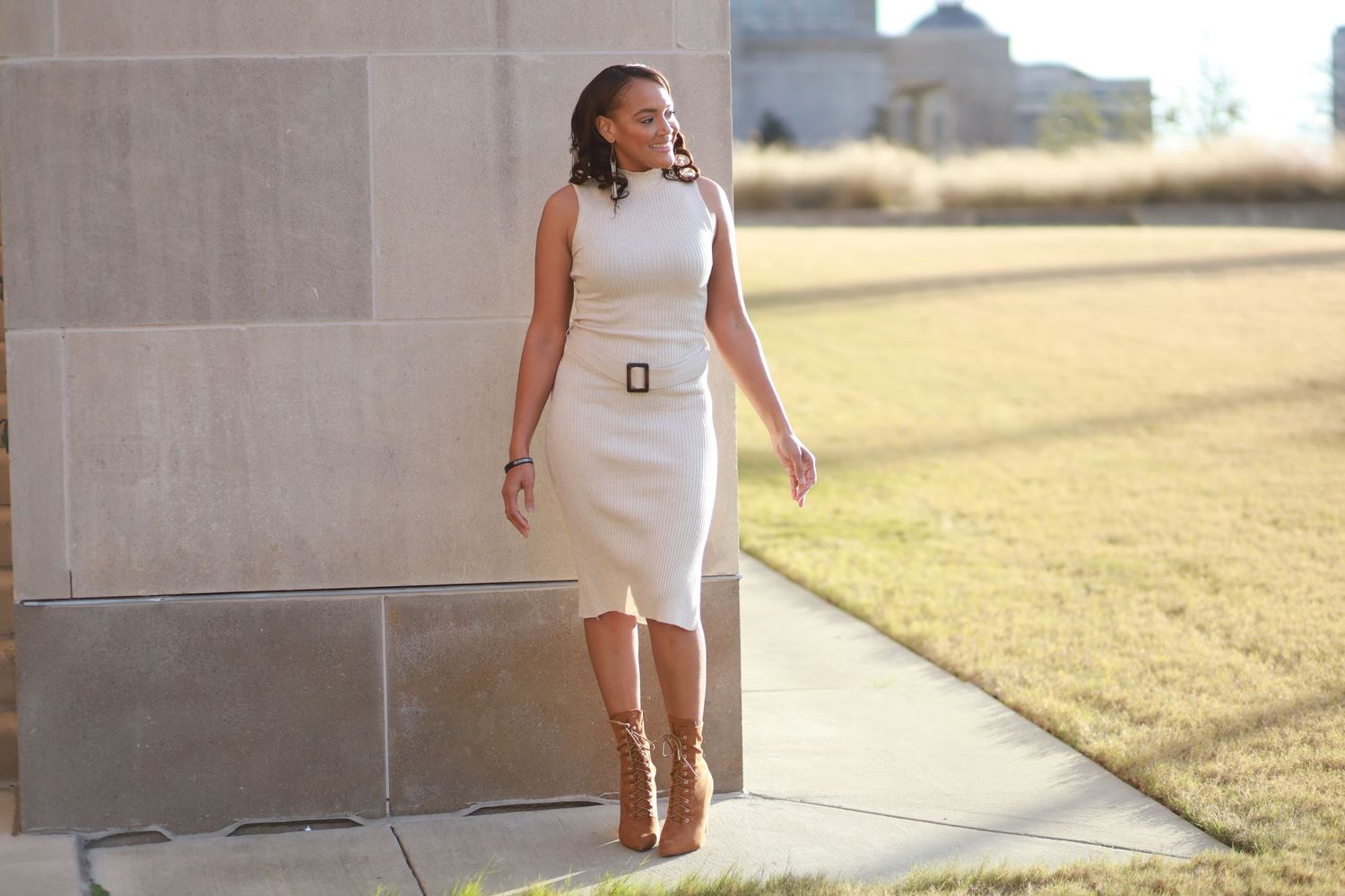 Woman in white fitted dress poses confidently against concrete wall outdoors.