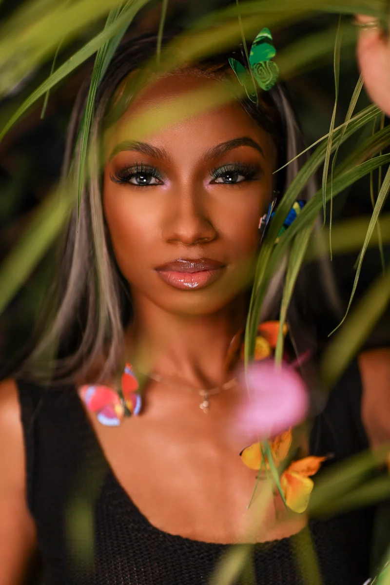 Artistic portrait with tropical leaves and butterflies framing subject against dark background.