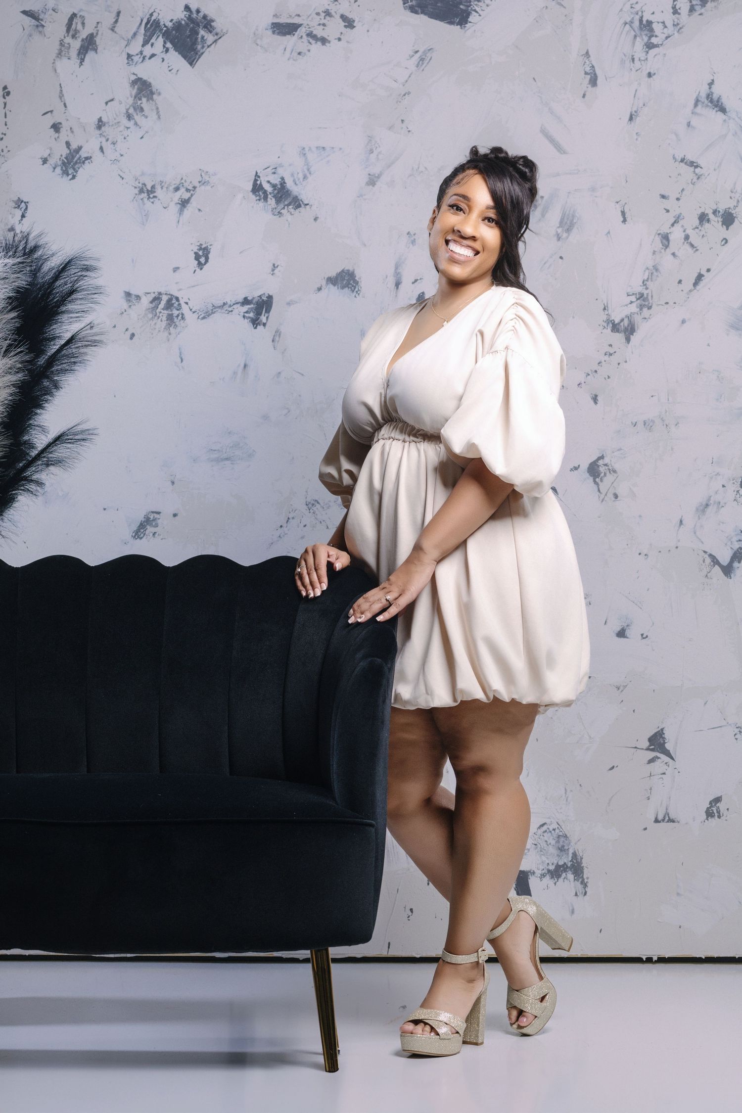 Woman in a cream-colored mini dress poses confidently next to a black sofa against a textured wall.