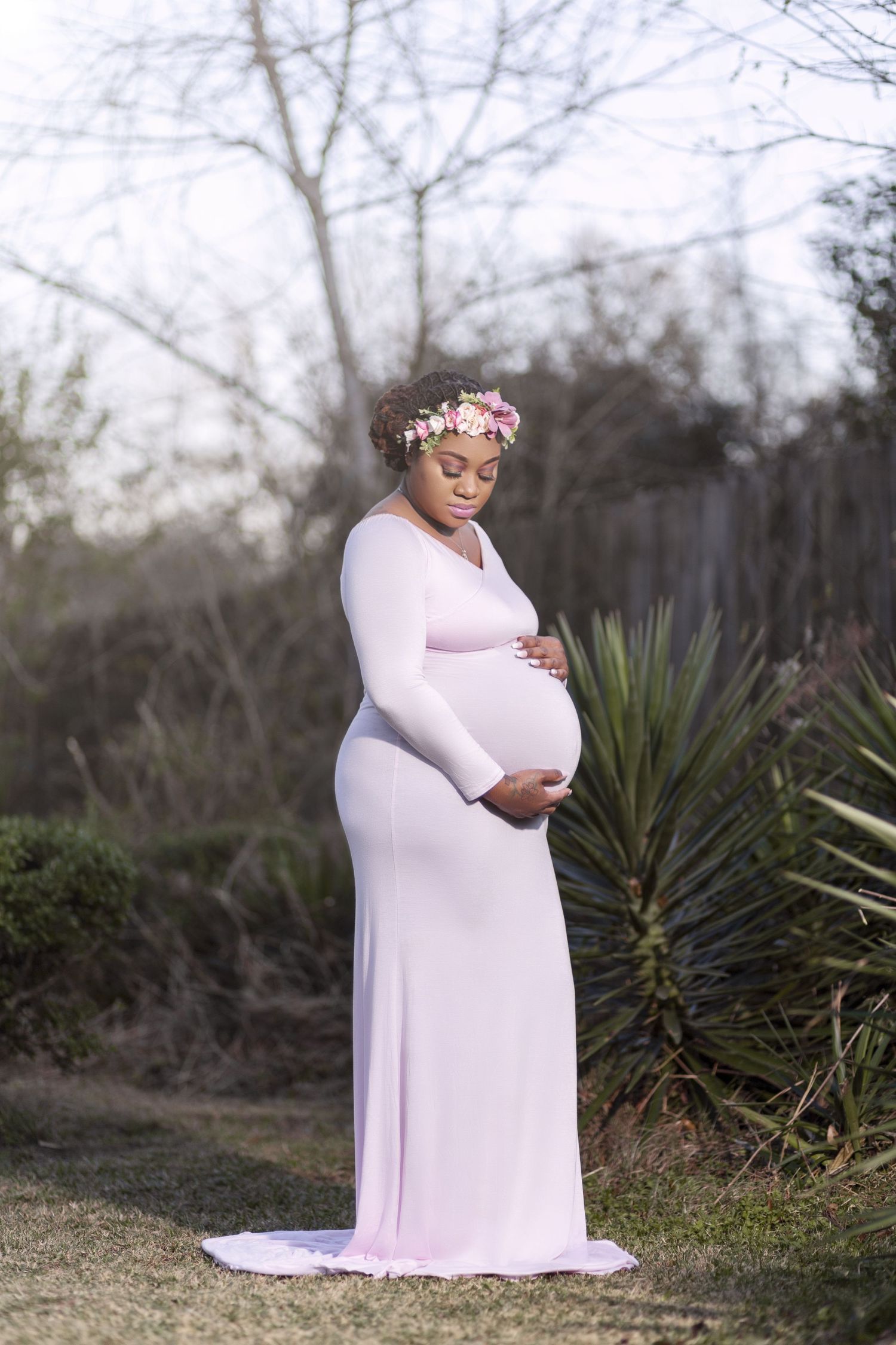 Maternity photo in pale pink dress with flower crown against natural outdoor setting.