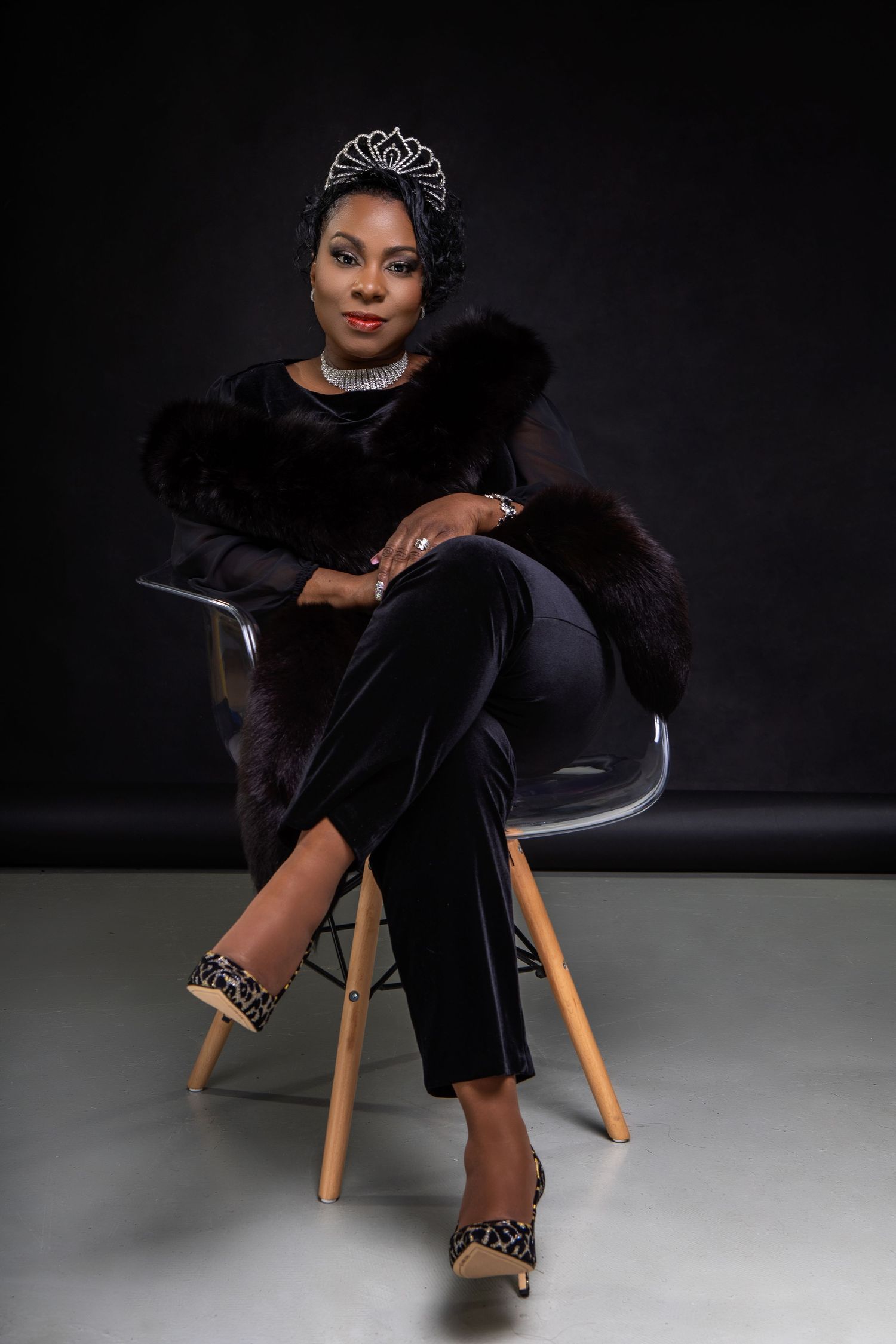 Elegant portrait of woman in black fur-trimmed outfit and tiara seated in modern chair.