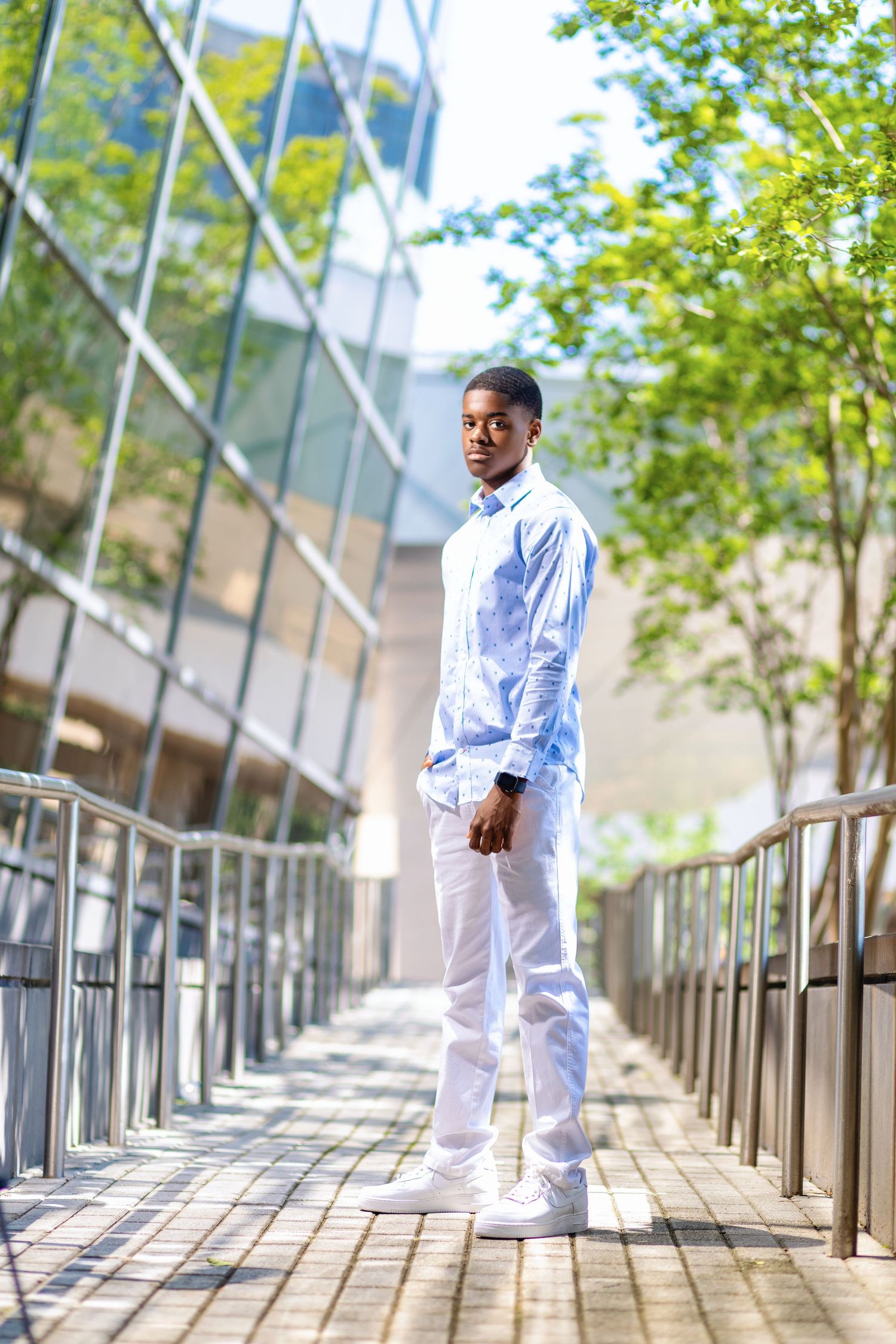 Modern urban fashion portrait on walkway with glass building and greenery.