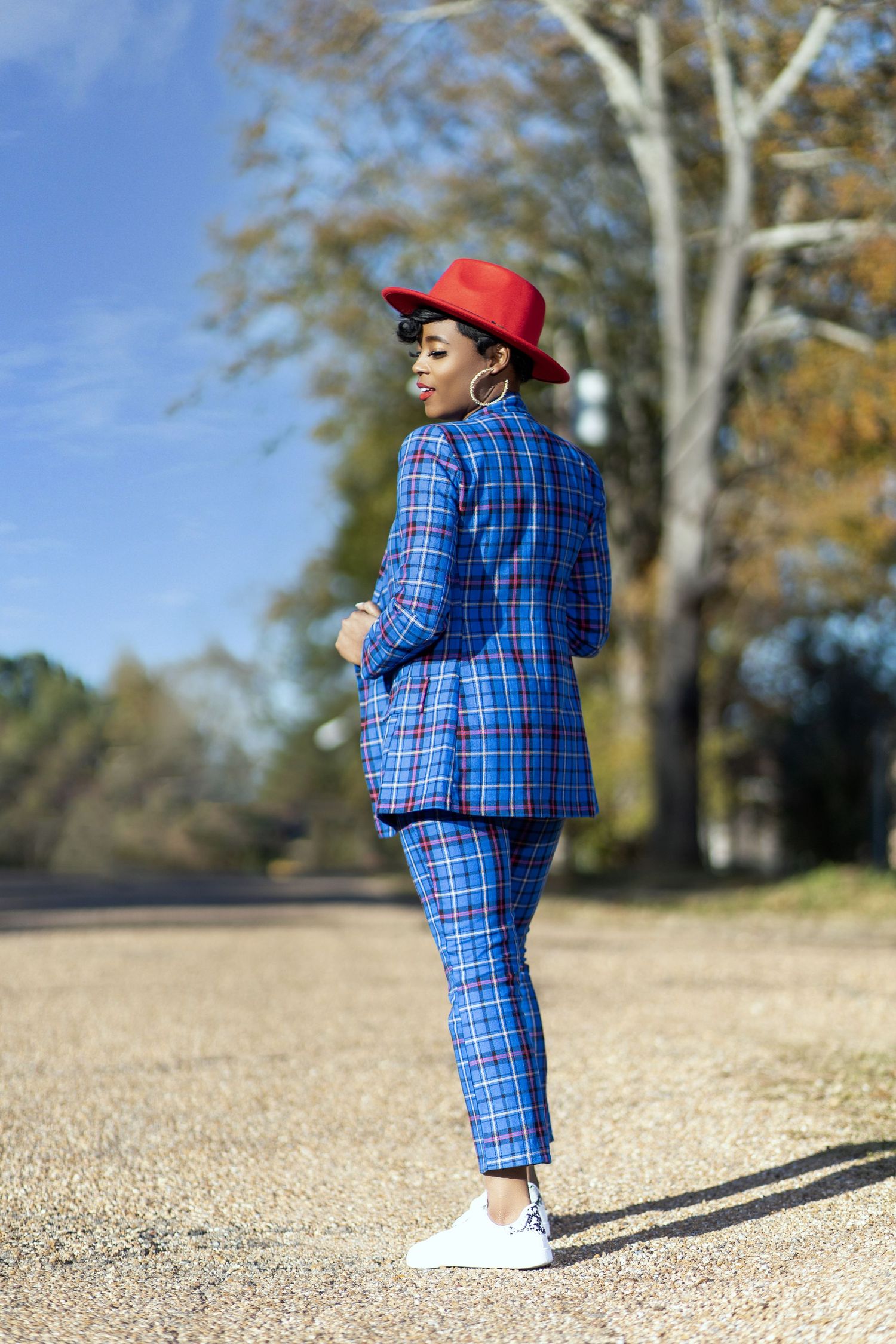 Person in bright blue plaid suit and red hat poses outdoors on dirt path during autumn.