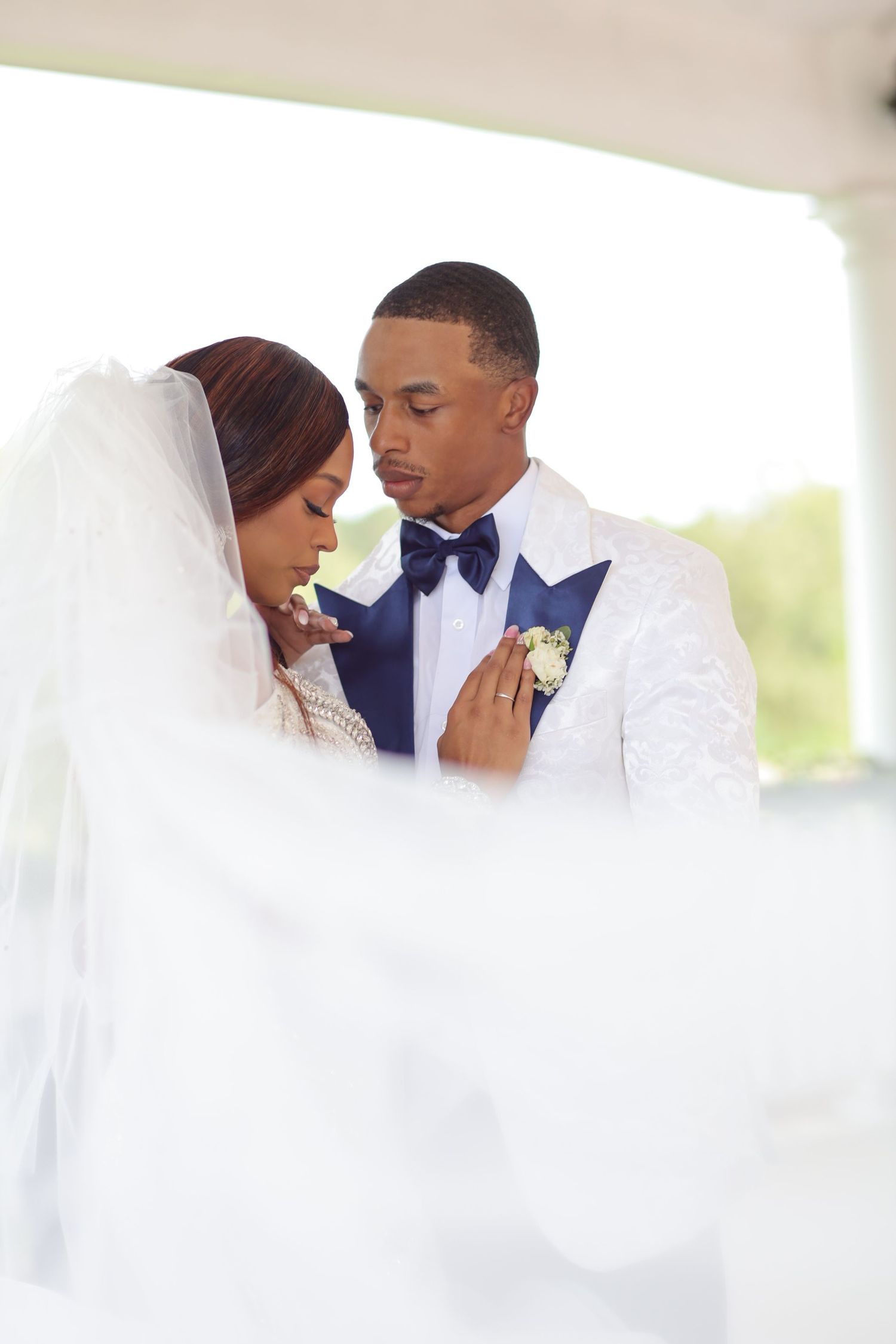 Wedding couple in elegant white attire share intimate moment under outdoor pavilion.
