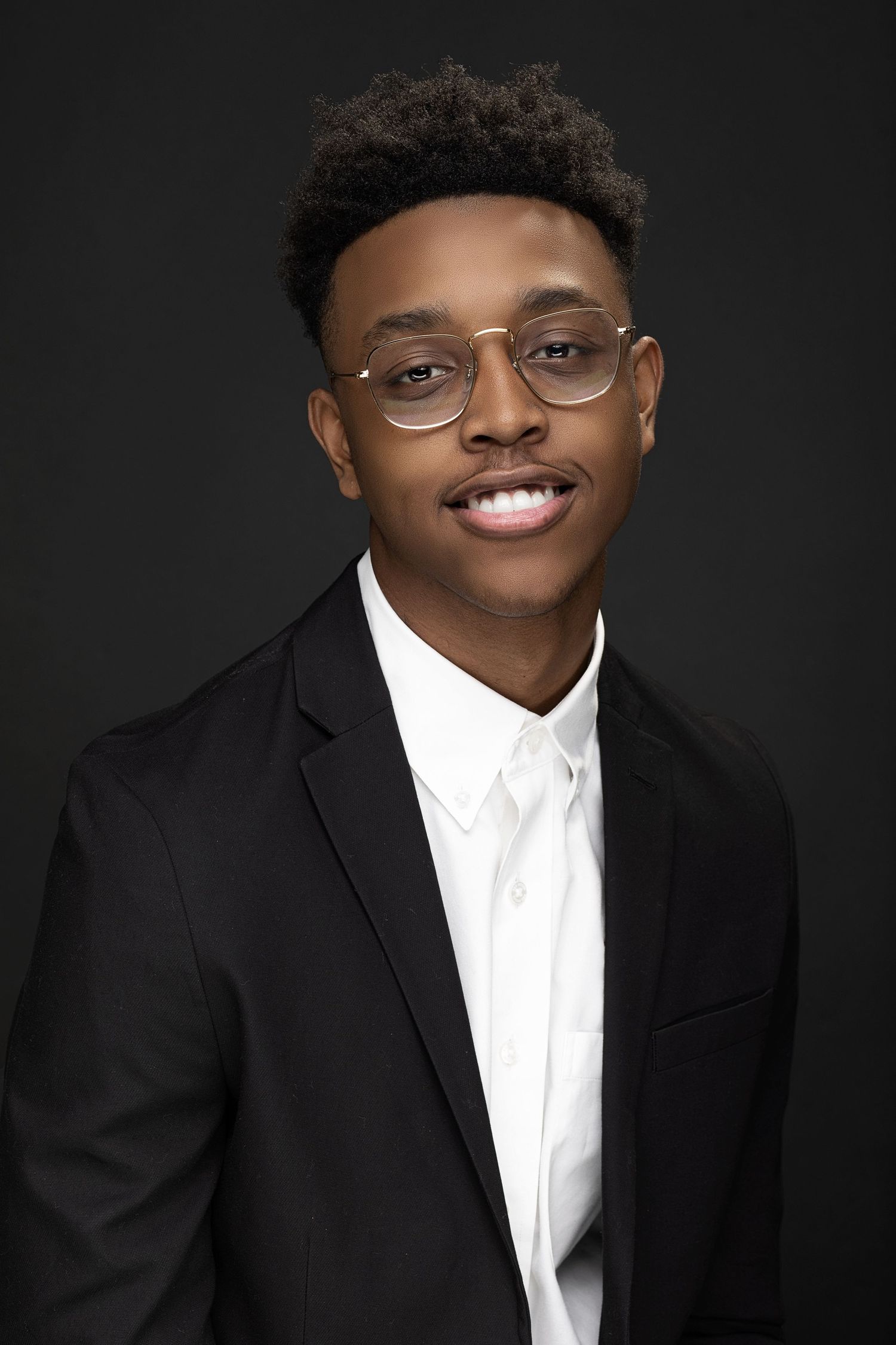 Professional headshot of young man in black suit and glasses against dark background.