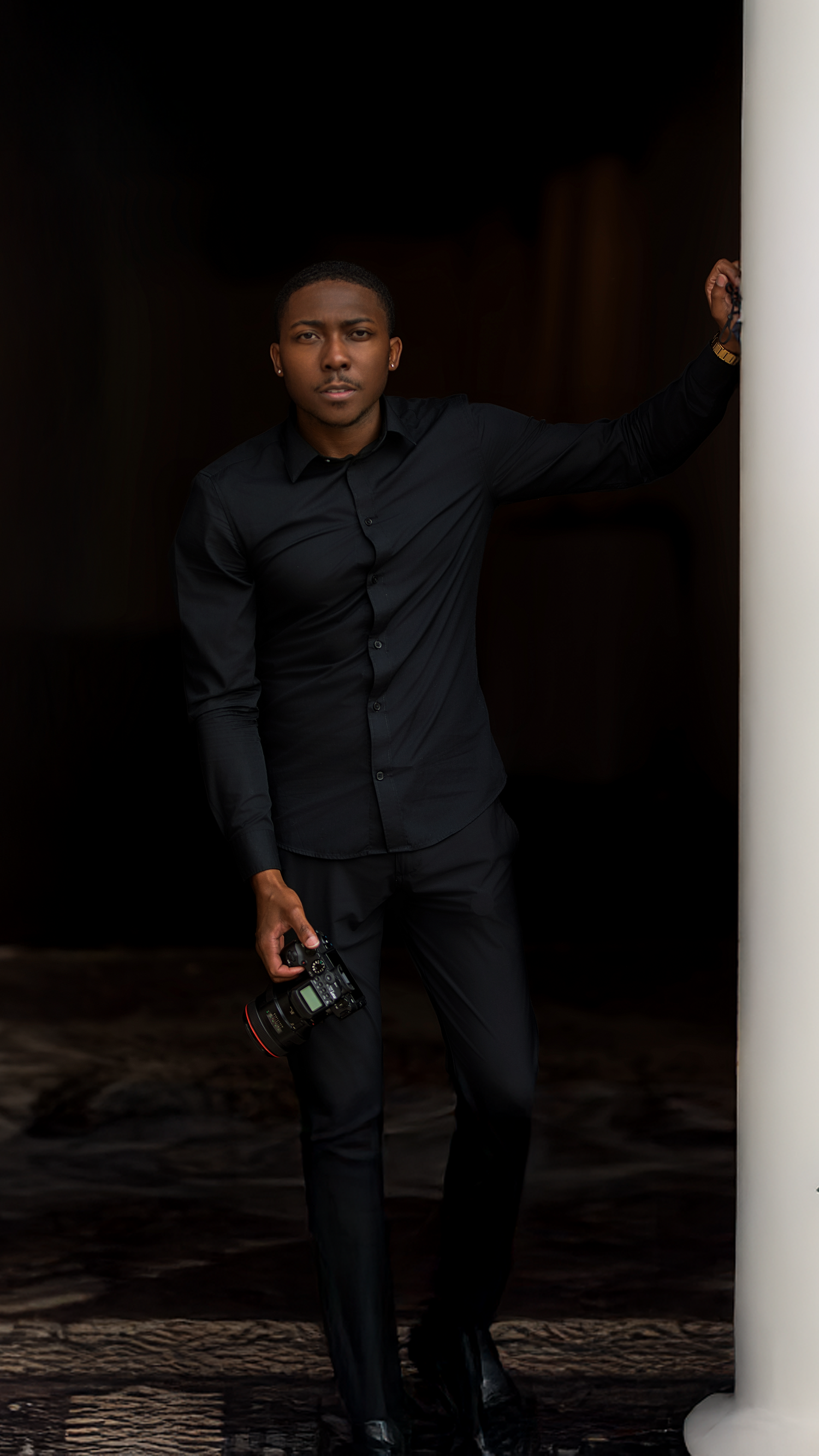 Professional photographer in black attire holding camera against dark backdrop.