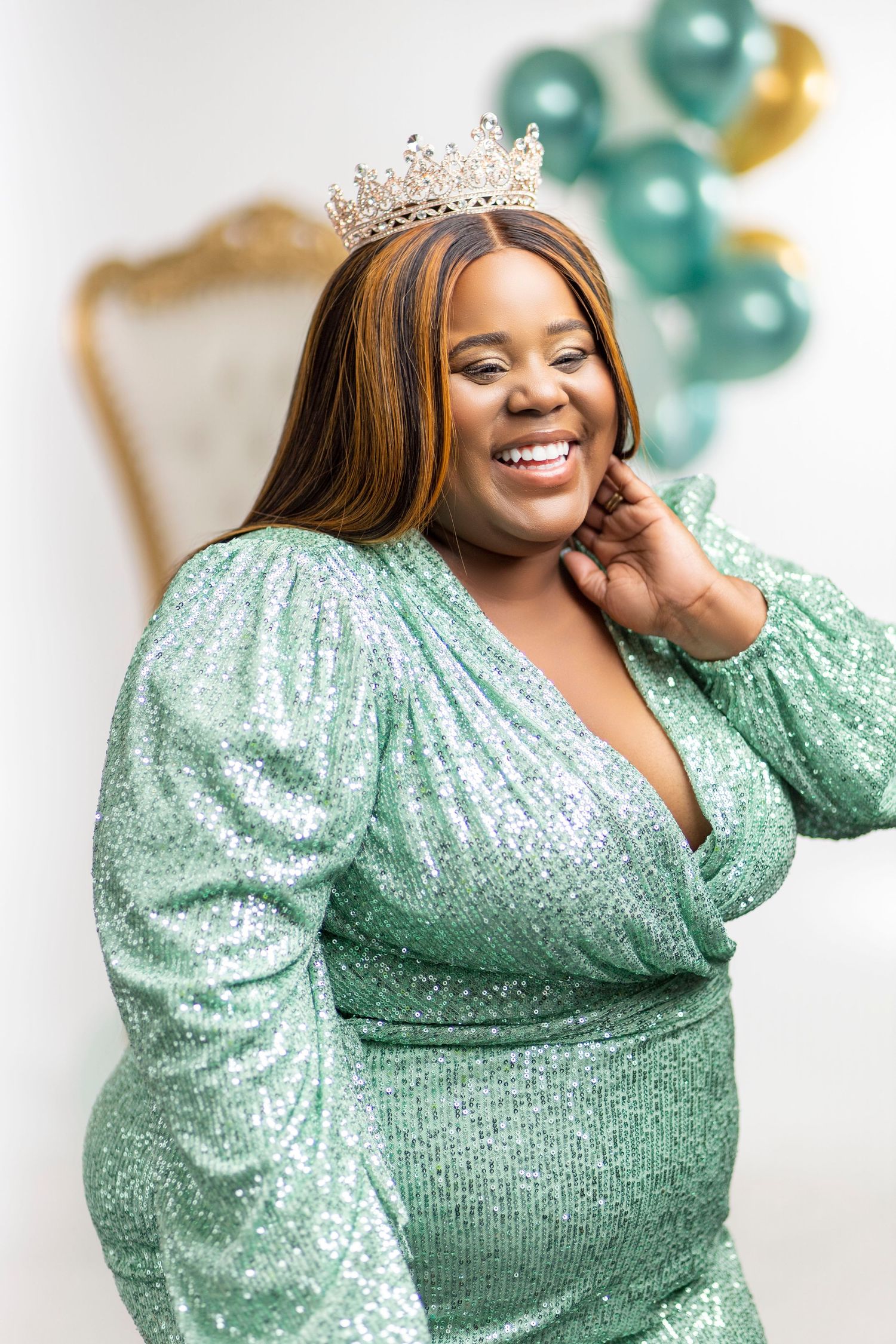 Celebratory portrait in a sparkly mint green dress with a crown against balloon decorations.