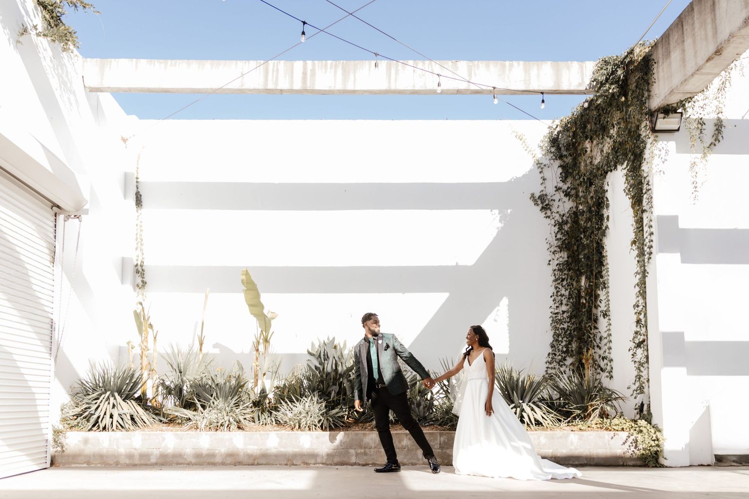 Newlywed couple dances in modern white architectural space with hanging plants.