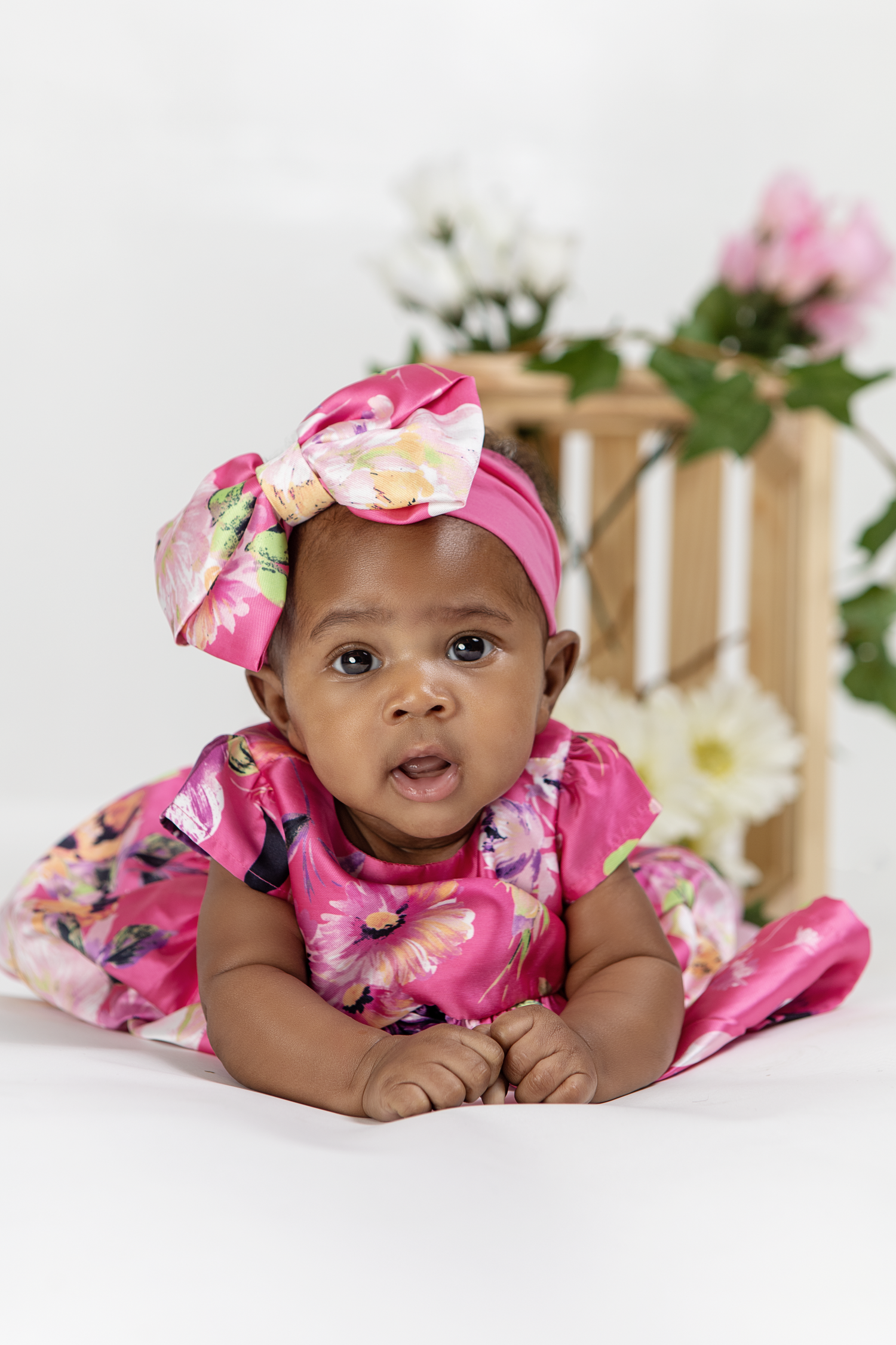Baby in pink floral dress and matching bow headband lying on white background with flowers.