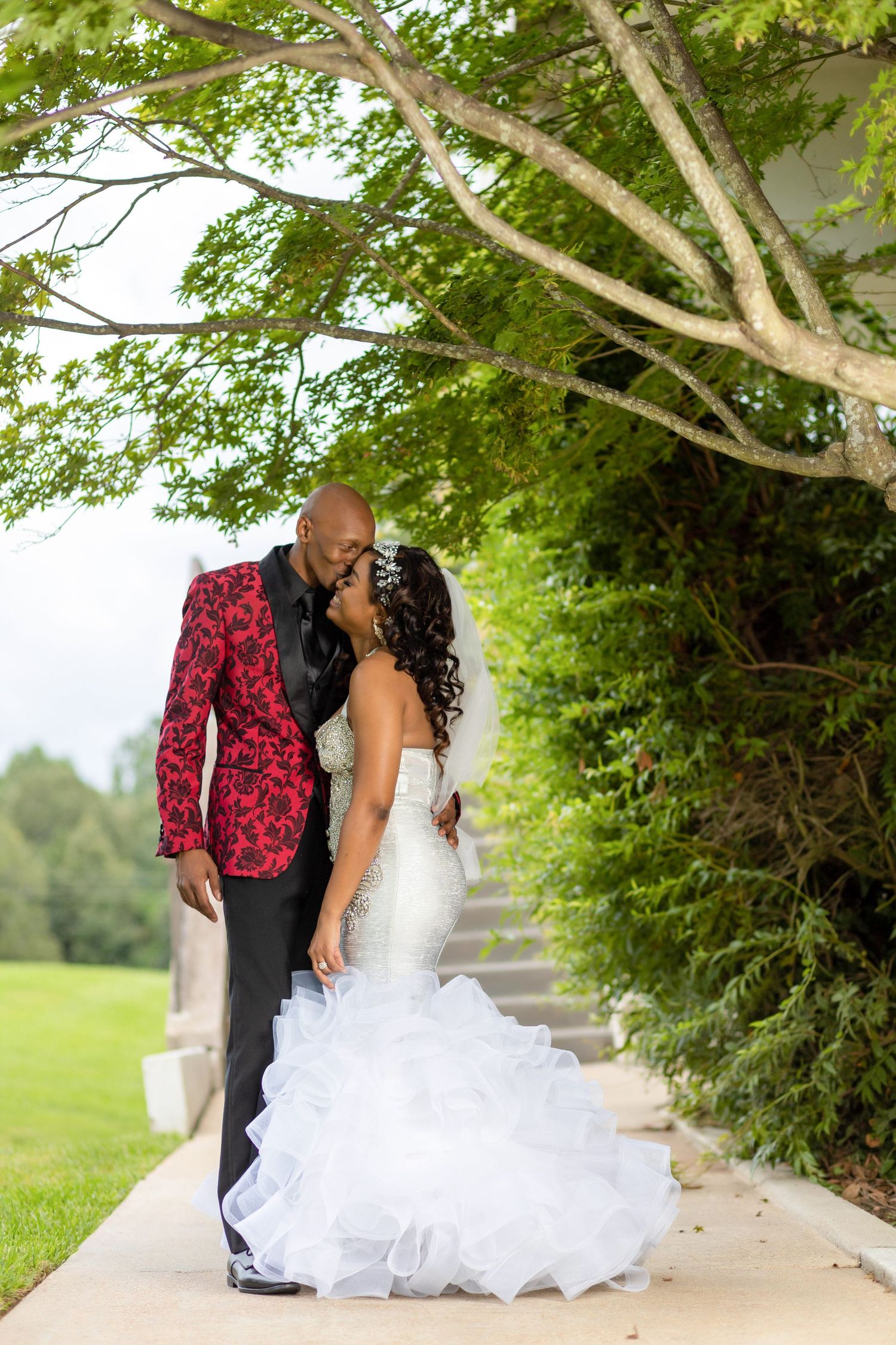 Romantic wedding photo of couple in red blazer and white gown beneath green trees.