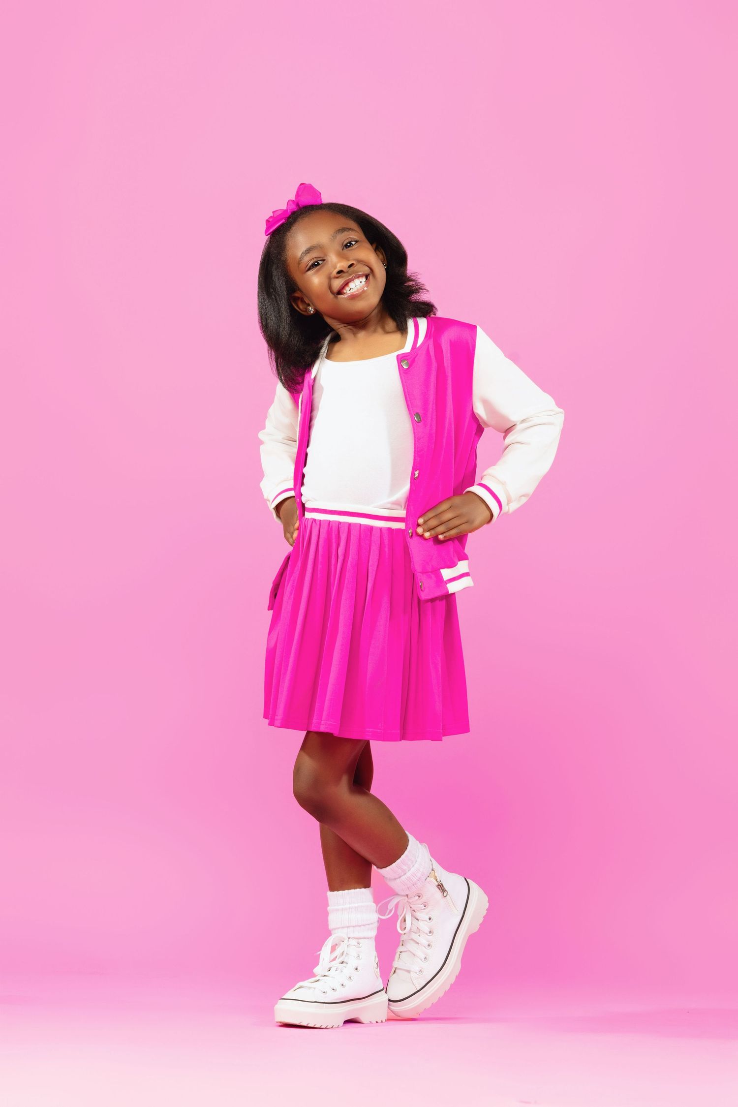 A child wearing a bright pink dress with white sleeves and matching bow poses cheerfully against a pink background.