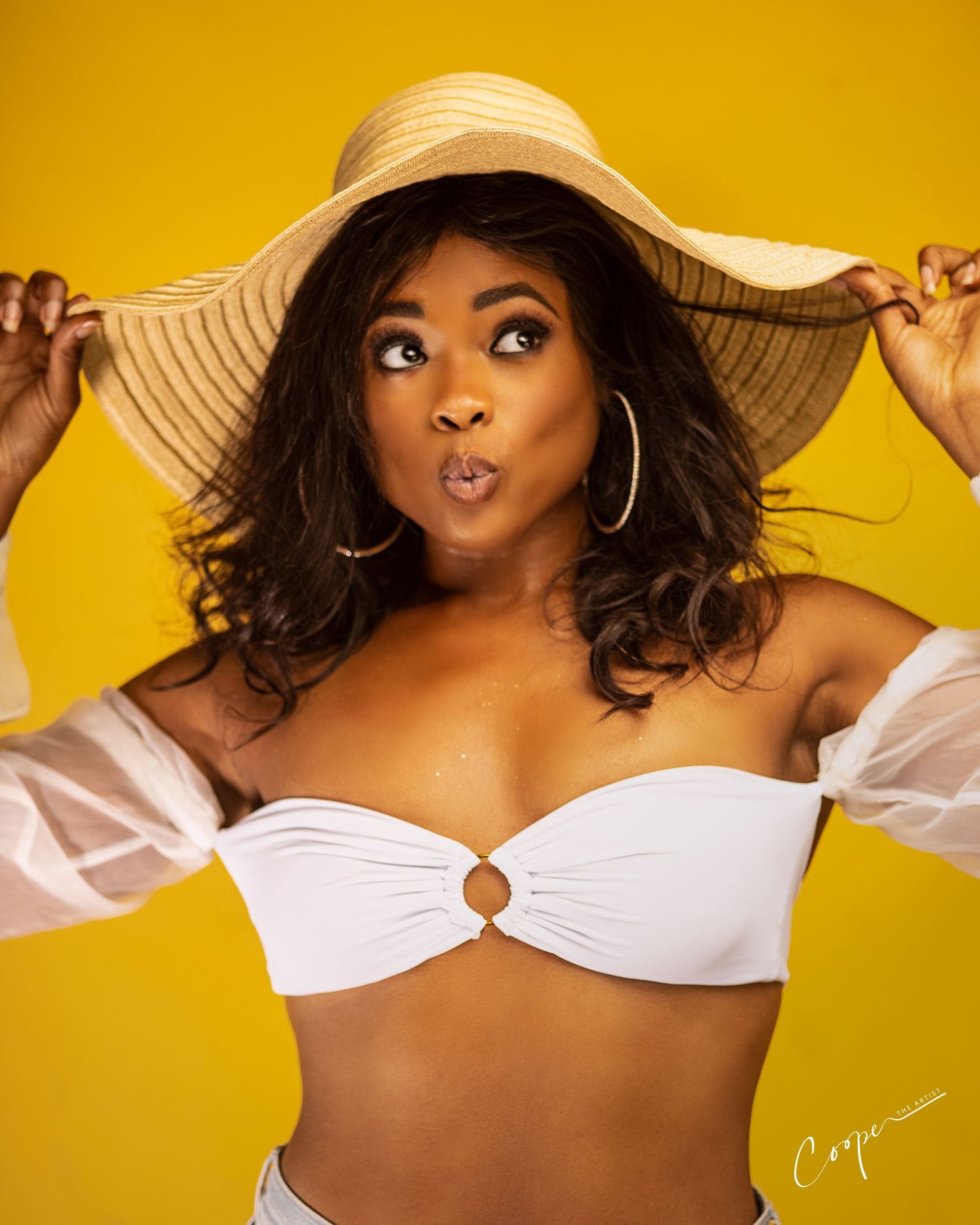 Fun summer portrait with playful expression and wide-brimmed hat against yellow background.