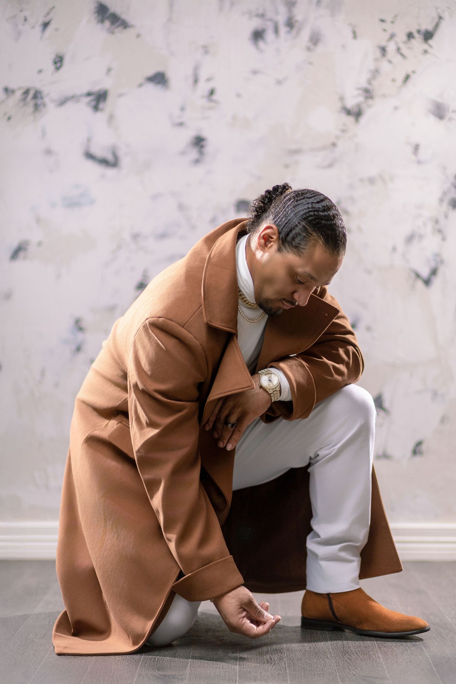 A person in a camel coat and white pants poses thoughtfully against a textured gray wall.
