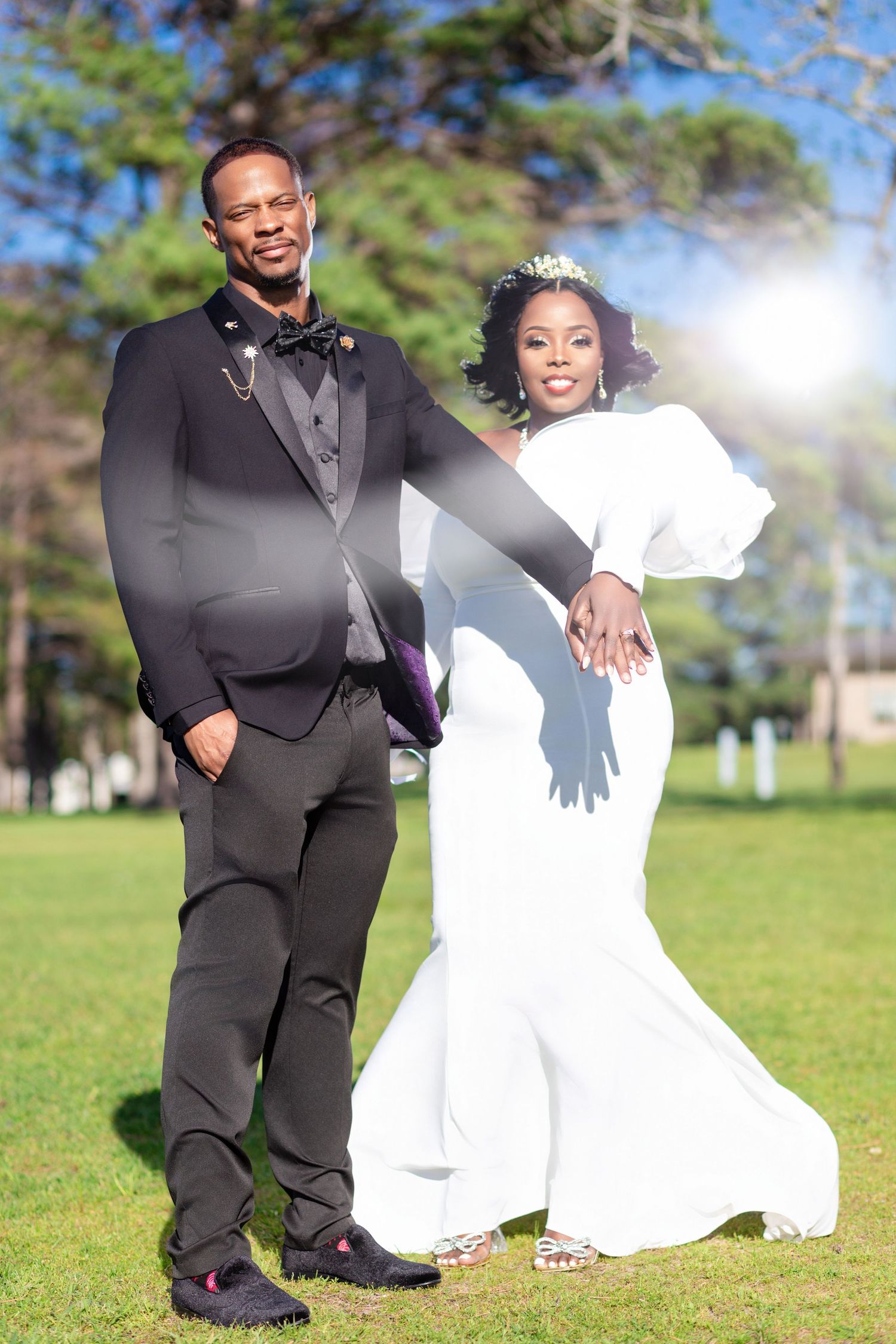 An elegant wedding couple posing outdoors in formal attire on a sunny day with trees in background.