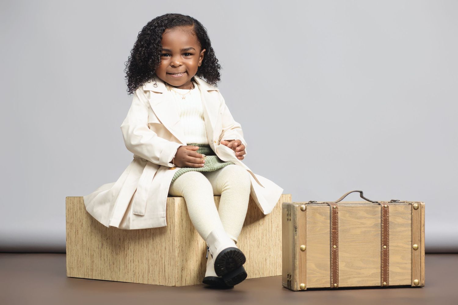 Cheerful child portrait in cream outfit sitting on wooden furniture against gray backdrop.