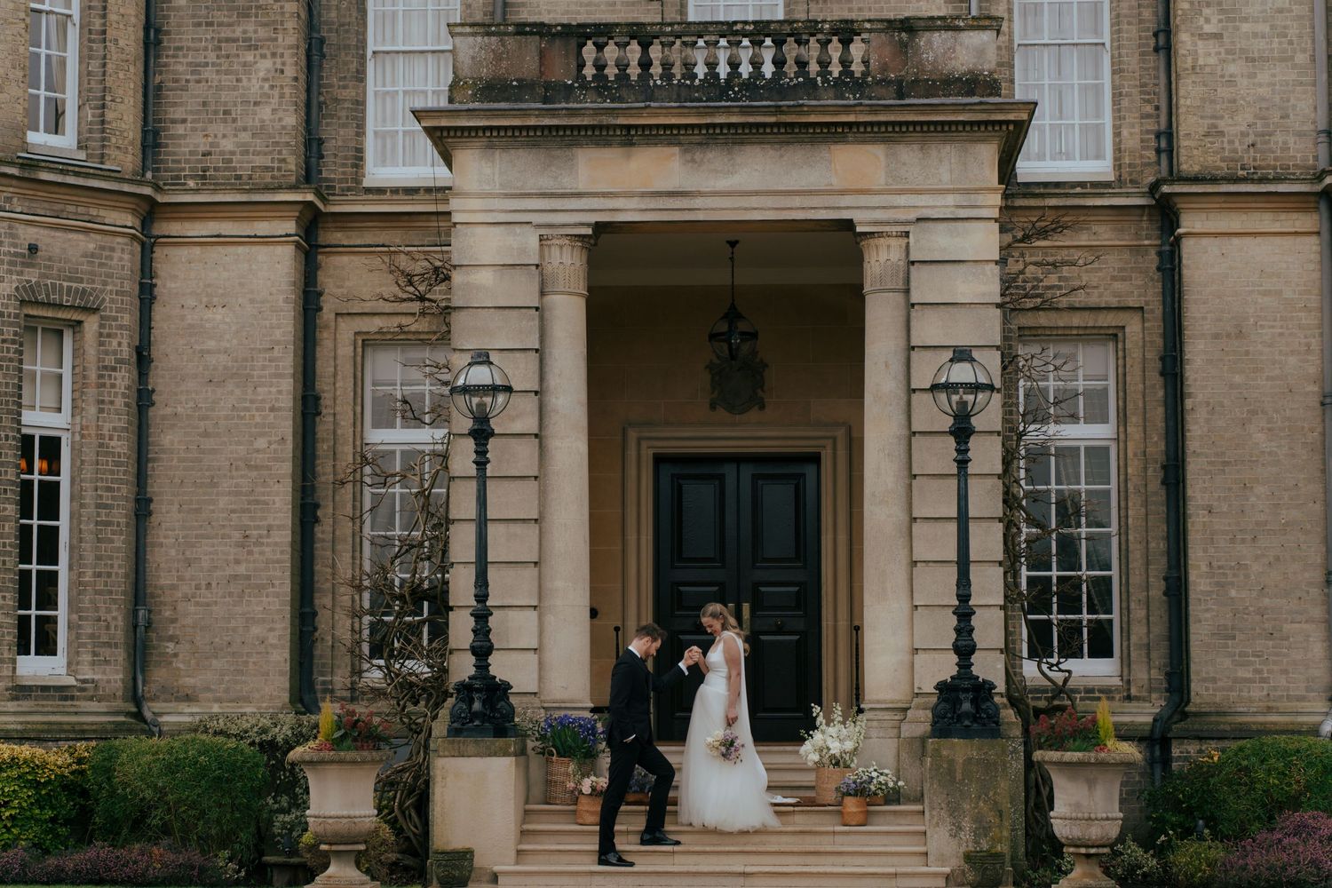 Two people share a tender moment on the grand stone steps of an elegant historic mansion with tall windows and ornate lanterns.