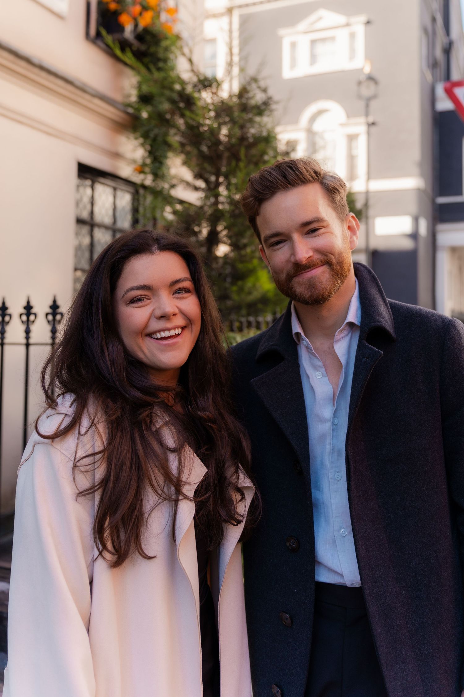 A smiling couple wearing light and dark coats pose together on a street with a white building behind them.