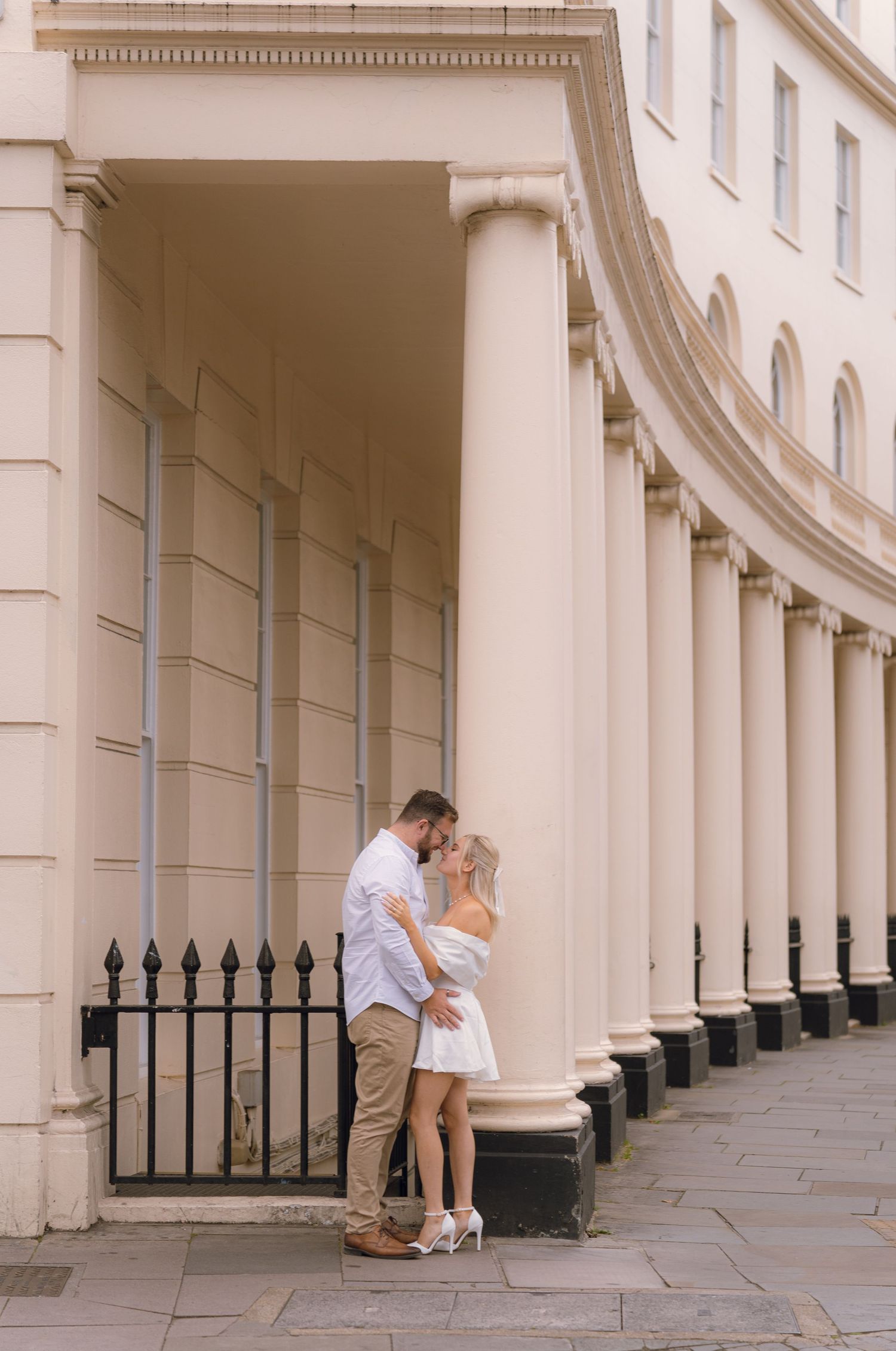 Romantic couple embracing against elegant curved colonnade with white columns and iron railings in London.