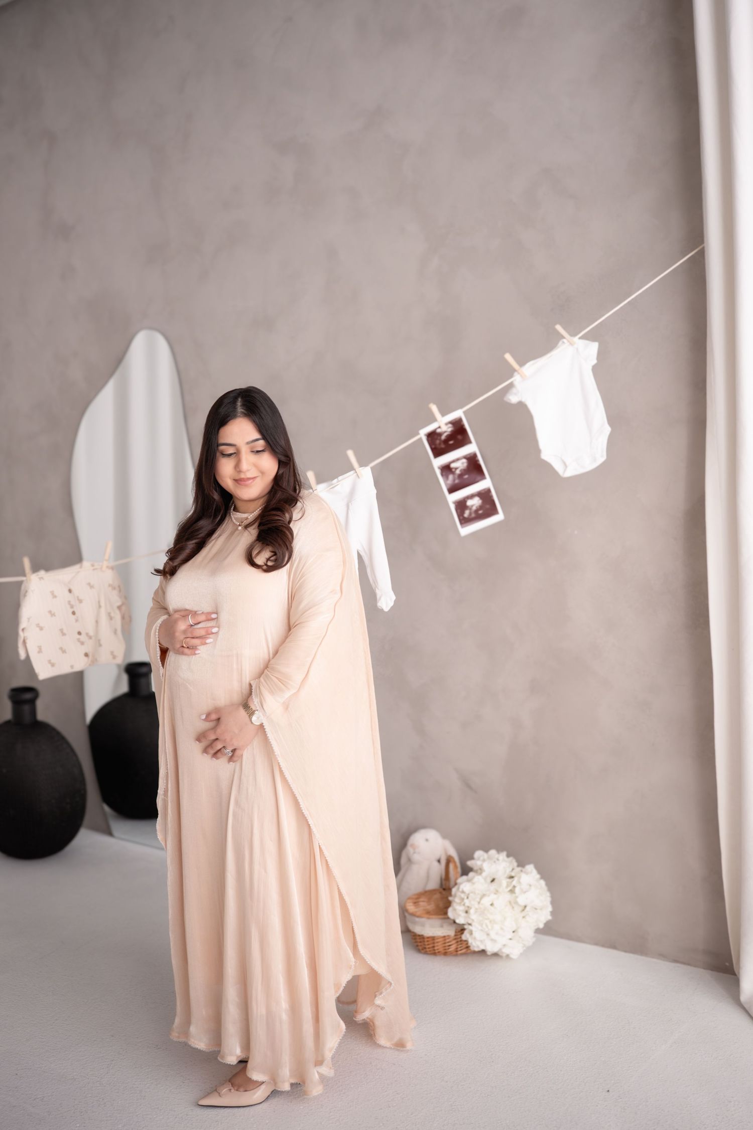 A pregnant woman in a flowing peach dress poses in a minimalist setting with baby clothes and ultrasound photos on a clothesline.